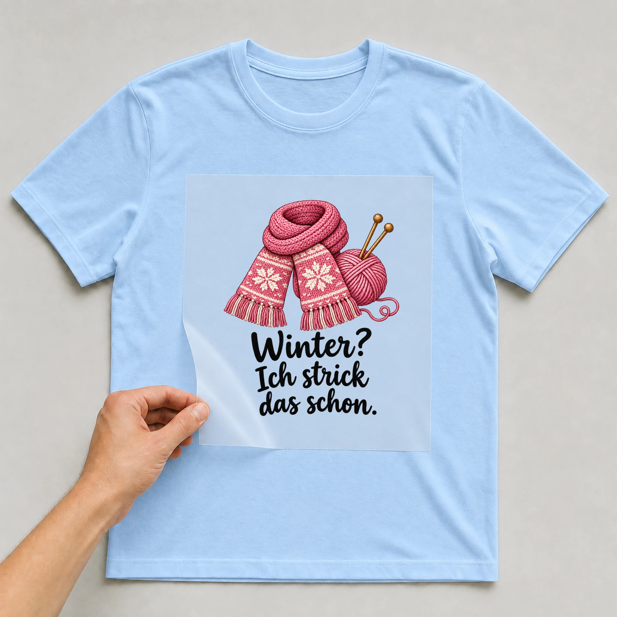 Bügelbild „Winter wird Masche für Masche“ auf babyblauem T-Shirt, Folie wird aufgelegt