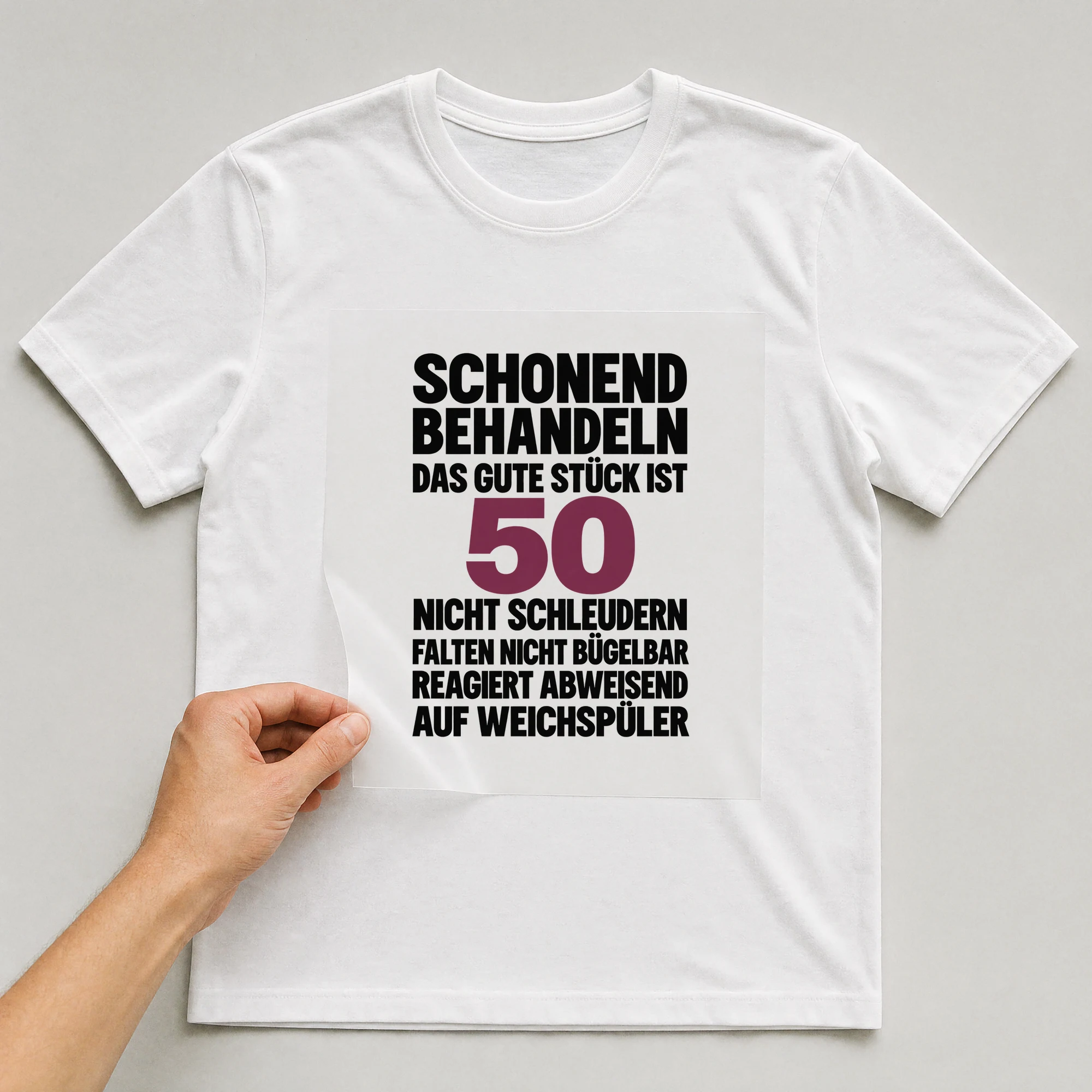 Bügelbild „Schonend behandeln das gute Stück ist 50 nicht schleudern falten nicht“ auf weißem T-Shirt, Folie wird aufgelegt