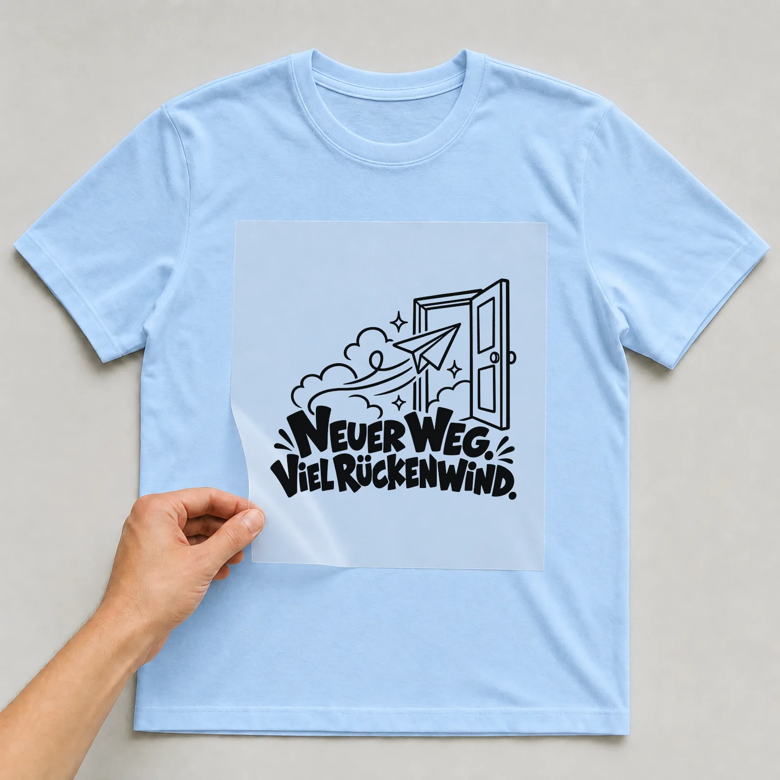 Bügelbild „Neuer weg. Viel Rückenwind.“ auf hellblauem T-Shirt, Folie wird aufgelegt