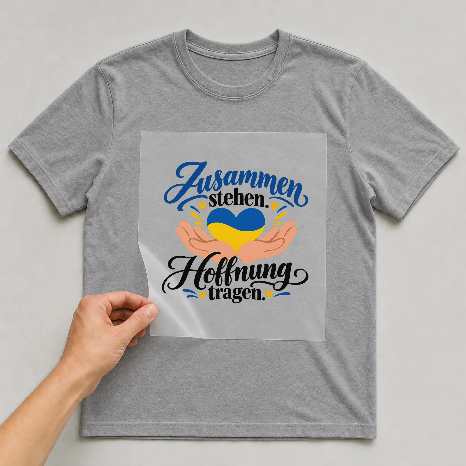 Bügelbild „Zusammen stehen. Hoffnung tragen.“ auf grauem T-Shirt, Folie wird aufgelegt
