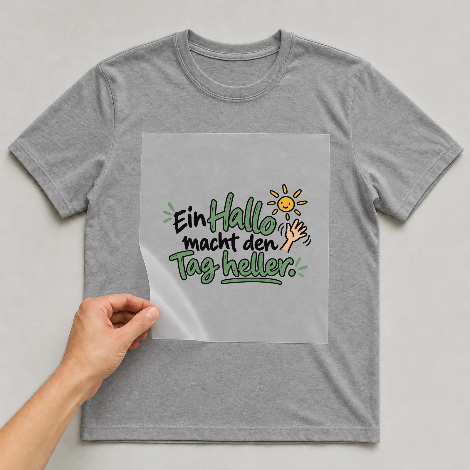 Bügelbild „Ein Hallo macht den Tag heller“ auf grauem T-Shirt, Folie wird aufgelegt