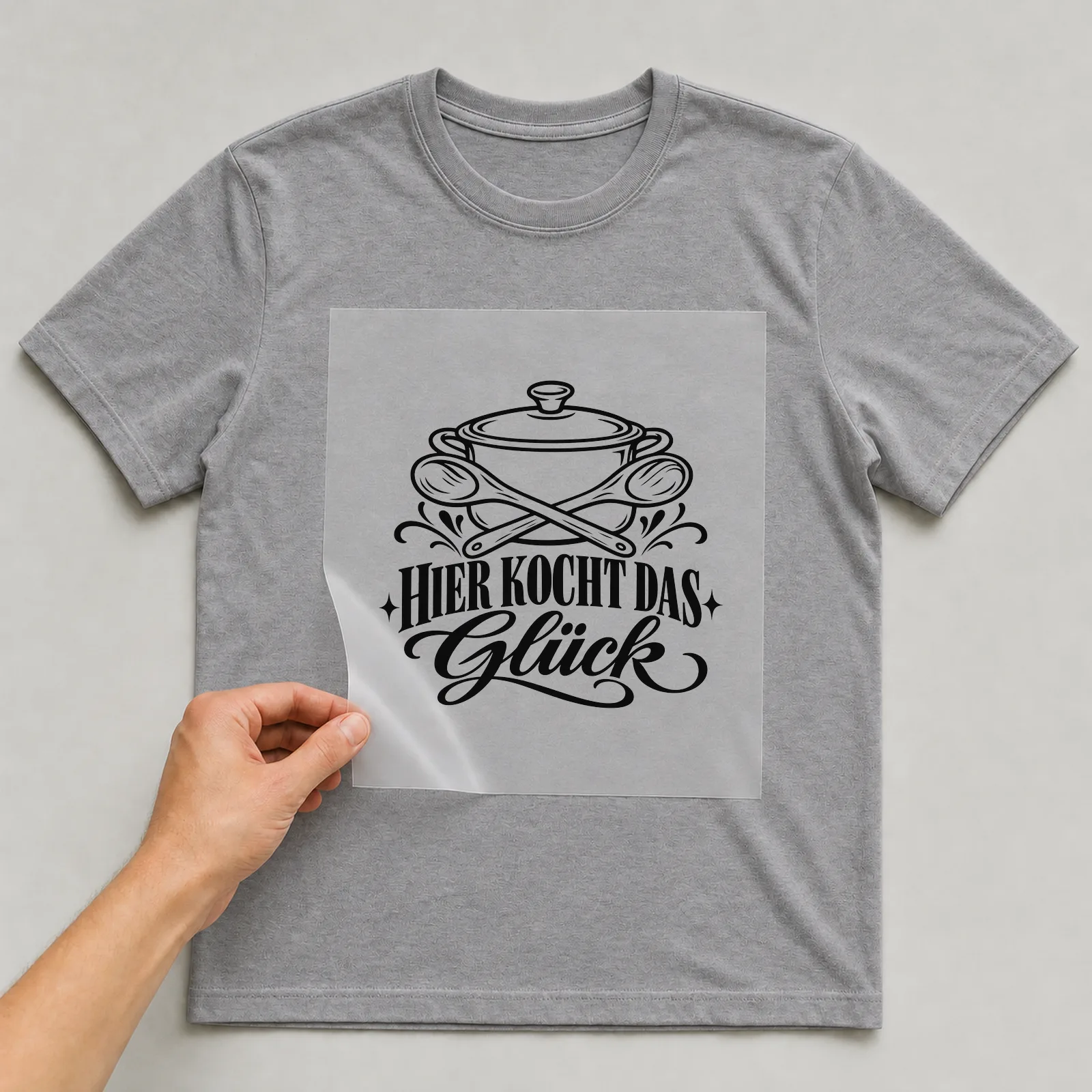 Bügelbild „Hier kocht das Glück“ auf grauem T-Shirt, Folie wird aufgelegt