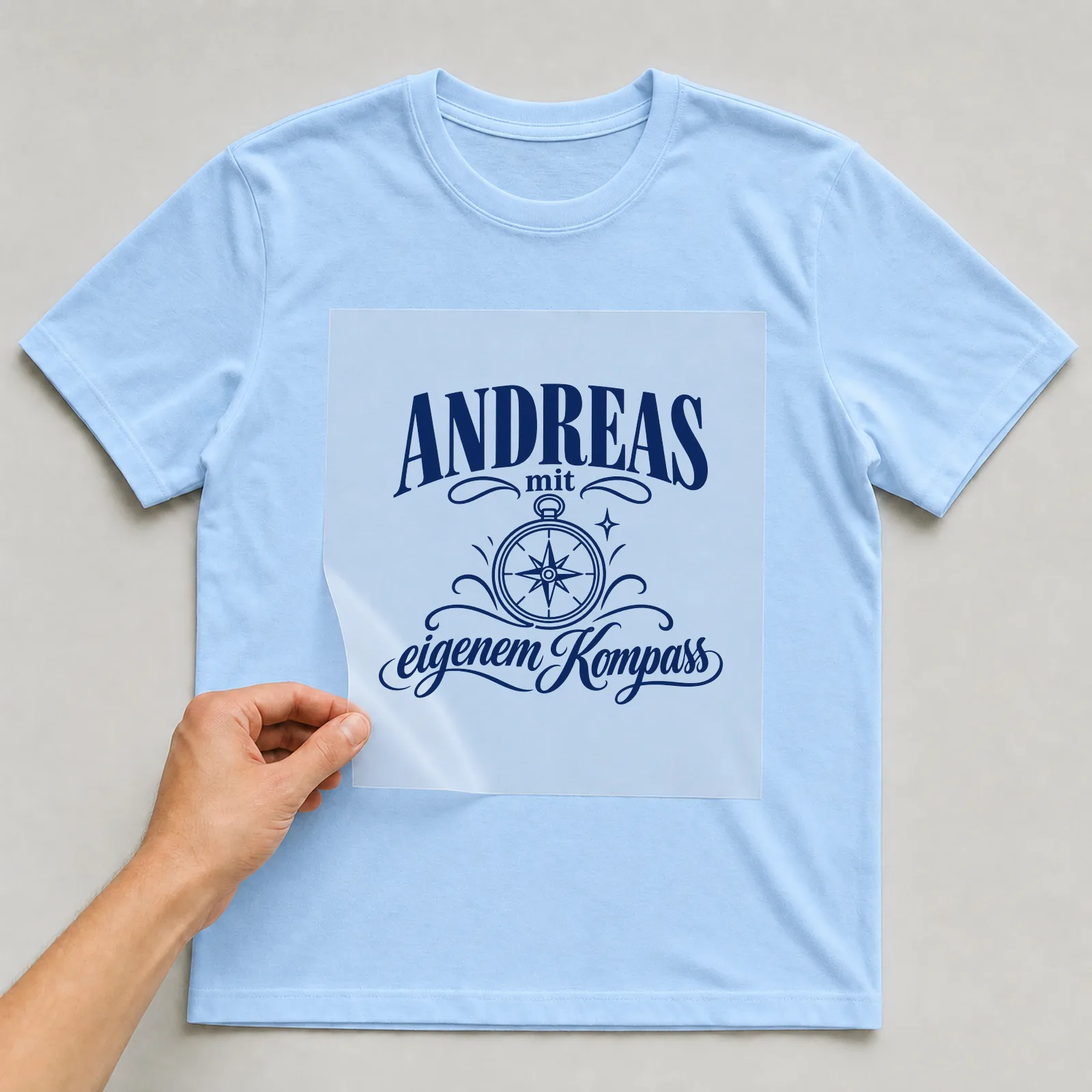 Bügelbild „Andreas mit eigenem Kompass“ auf hellblauem T-Shirt, Folie wird aufgelegt