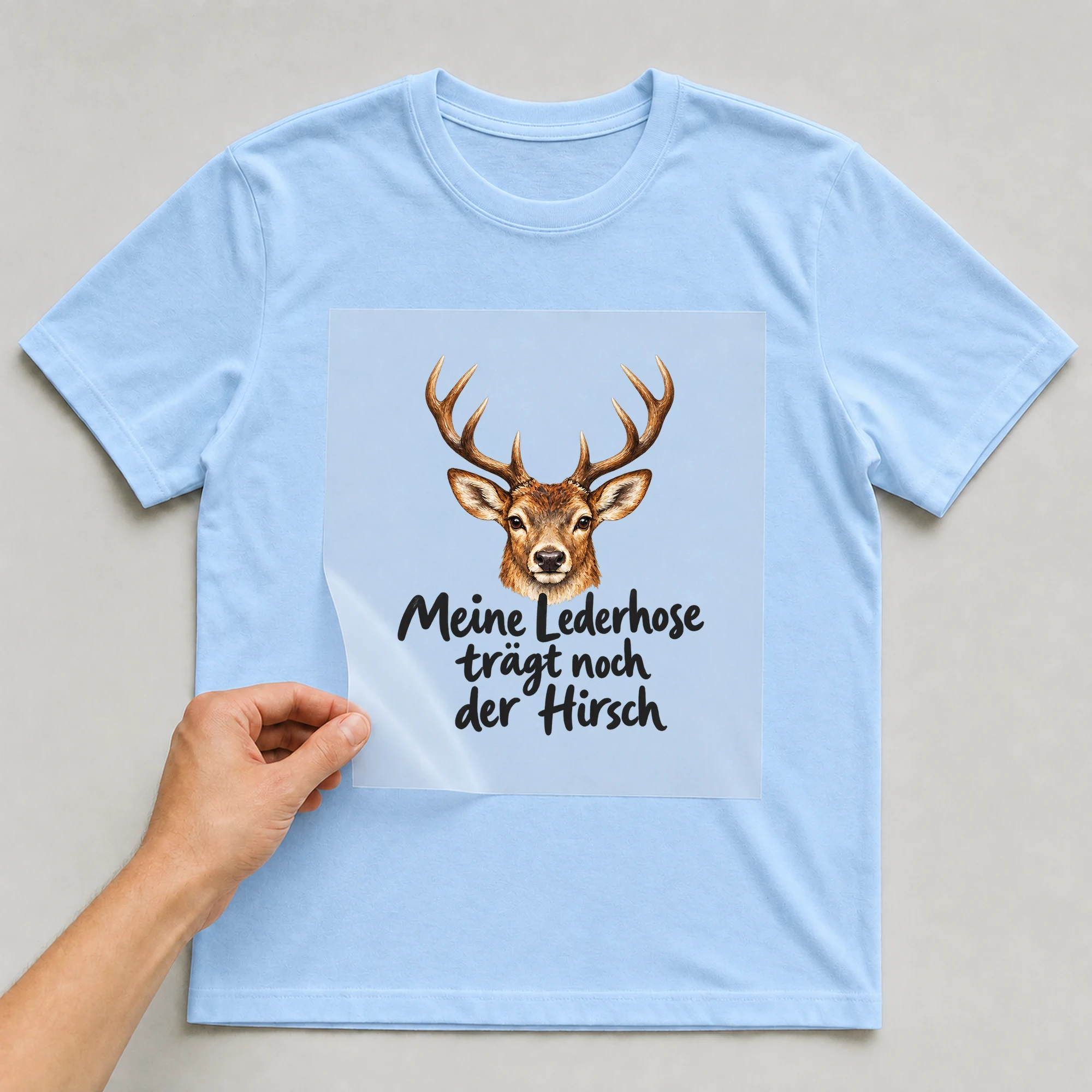 Bügelbild „Meine Lederhose trägt noch der Hirsch“ auf babyblauem T-Shirt, Folie wird aufgelegt