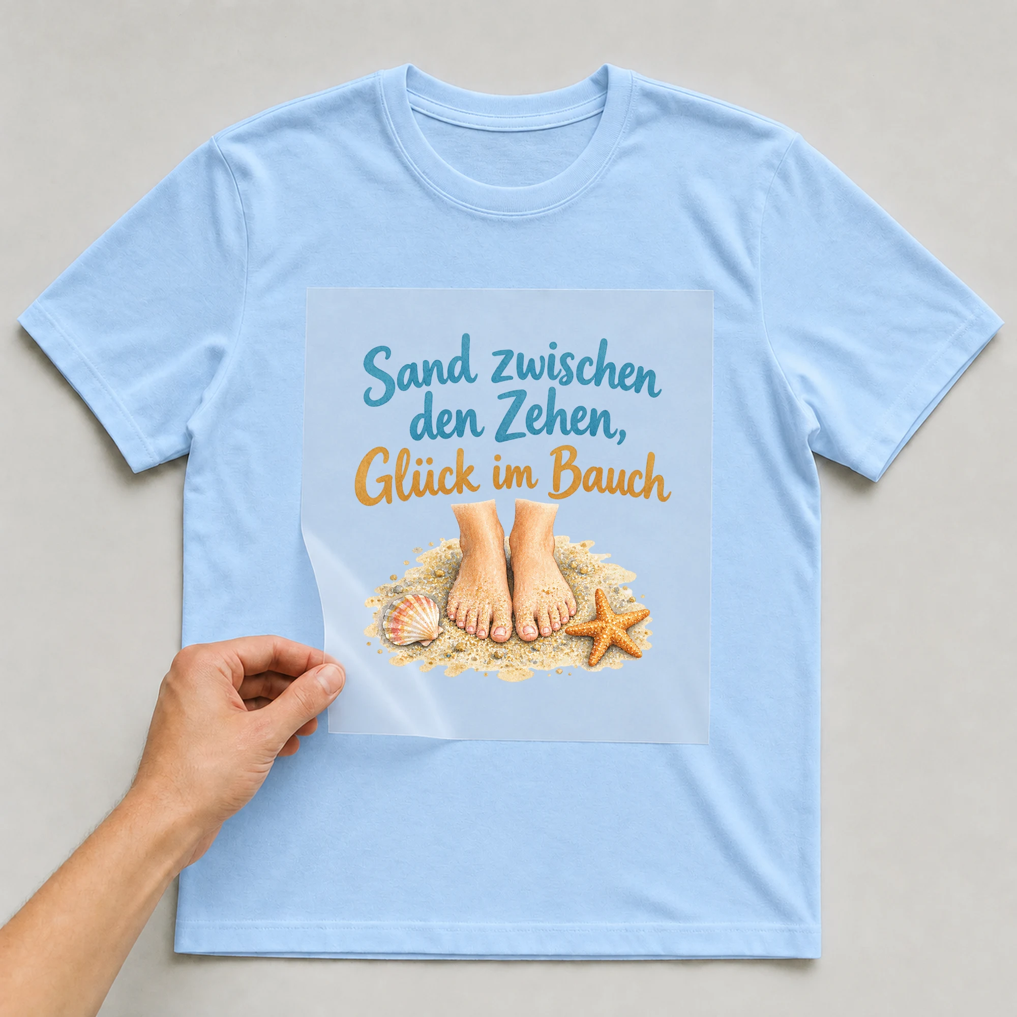 Bügelbild „Sand zwischen den Zehen, Glück im Bauch“ auf babyblauem T-Shirt, Folie wird aufgelegt