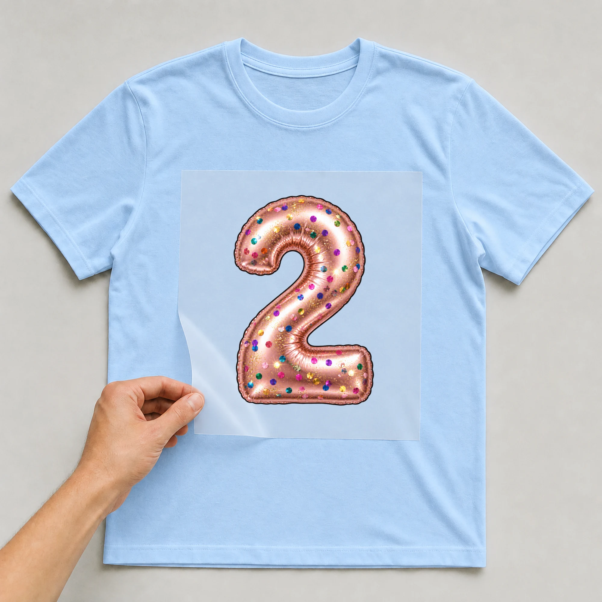 Bügelbild „Zahl 2 im Konfetti-Metallic-Look“ auf babyblauem T-Shirt, Folie wird aufgelegt