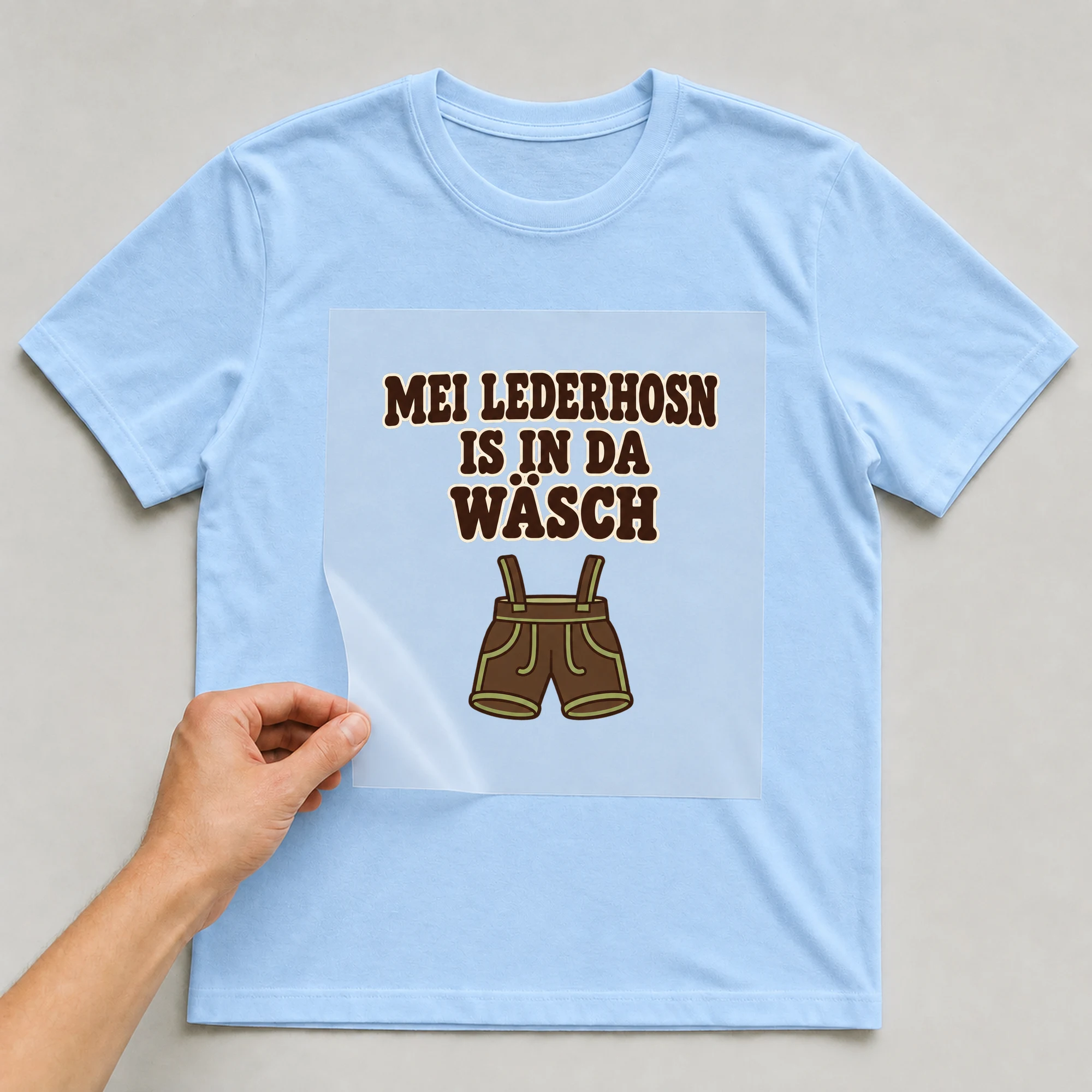 Bügelbild „MEI LEDERHOSN IS IN DA WÄSCH“ auf babyblauem T-Shirt, Folie wird aufgelegt