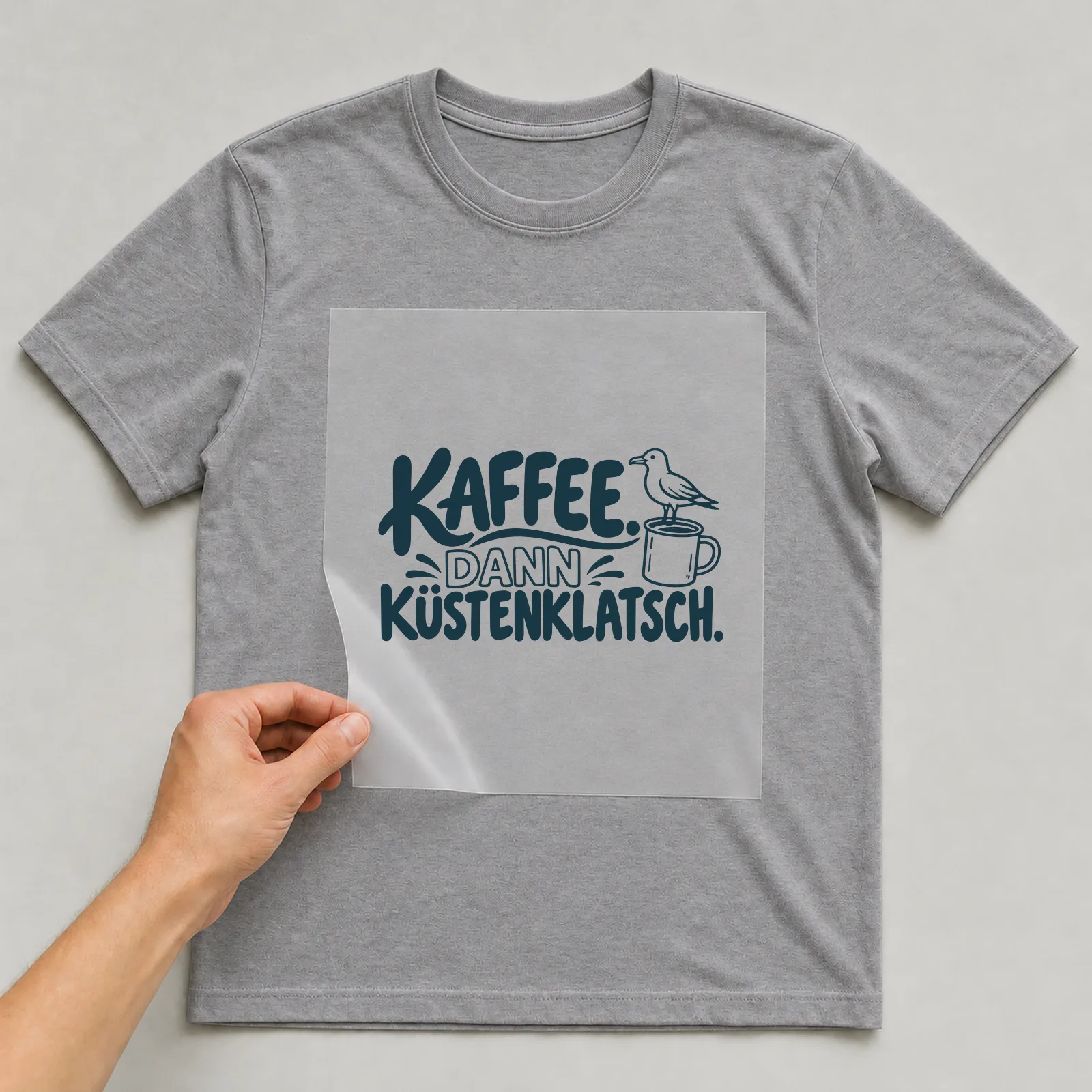 Bügelbild „Kaffee. Dann Küstenklatsch.“ auf grauem T-Shirt, Folie wird aufgelegt