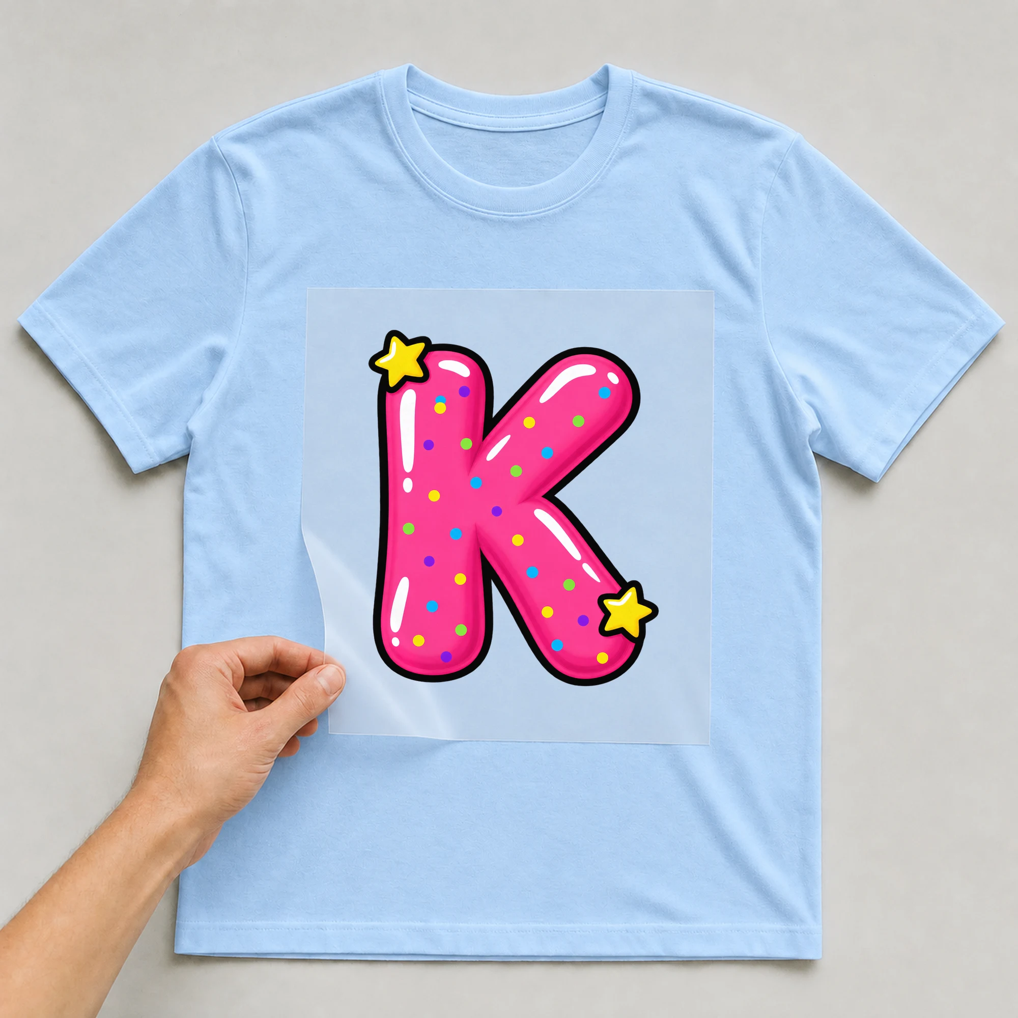 Bügelbild „Buchstabe K im Comic-Ballon-Stil“ auf babyblauem T-Shirt, Folie wird aufgelegt
