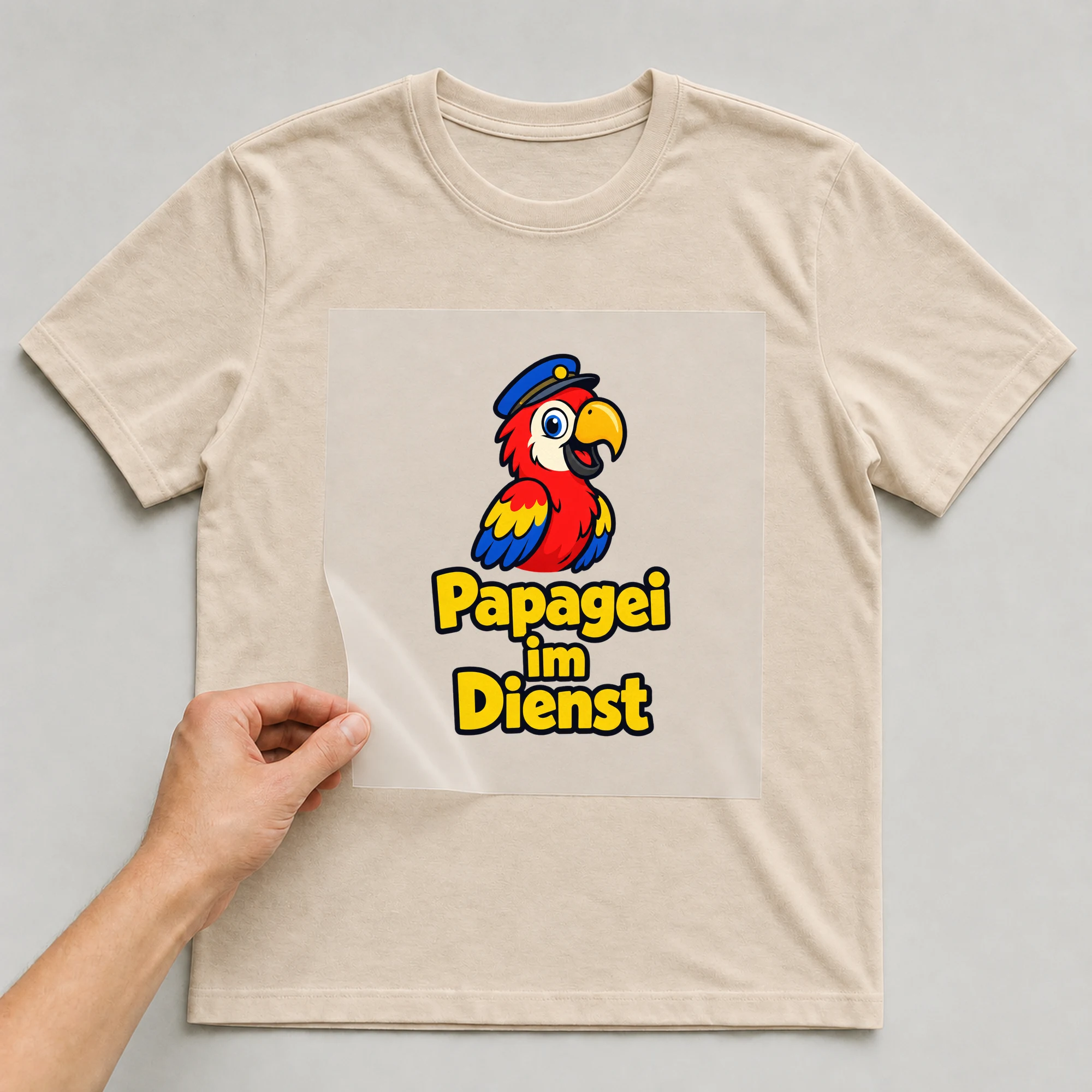 Bügelbild „Papagei im Dienst“ auf beigem T-Shirt, Folie wird aufgelegt