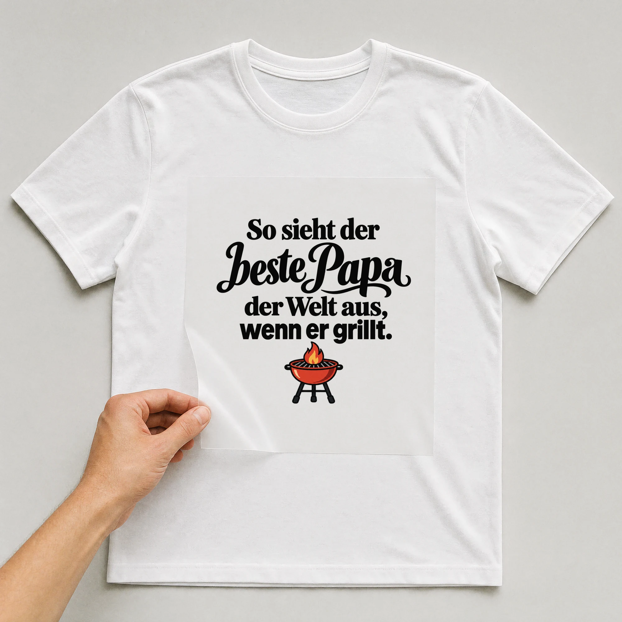 Bügelbild „So sieht der beste Papa der Welt aus, wenn er grillt.“ auf weißem T-Shirt, Folie wird aufgelegt