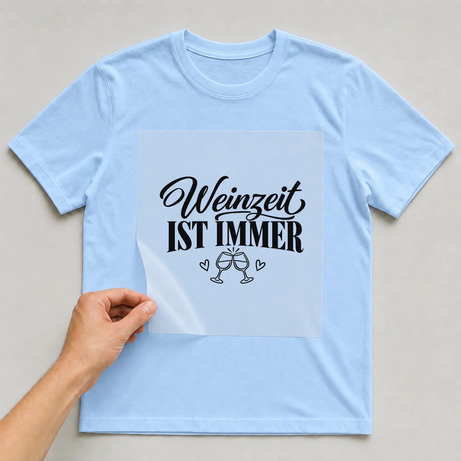 Bügelbild „Weinzeit ist immer“ auf hellblauem T-Shirt, Folie wird aufgelegt