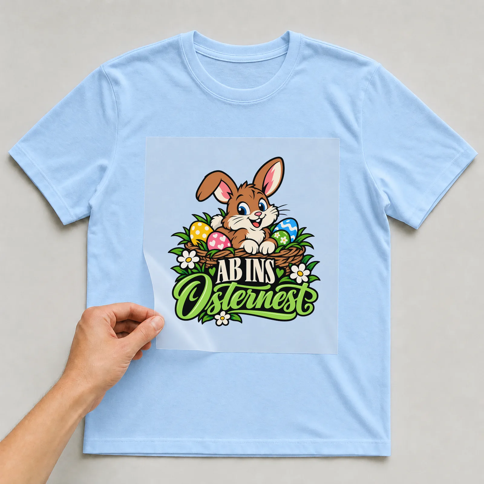 Bügelbild „Ab ins Osternest“ auf hellblauem T-Shirt, Folie wird aufgelegt