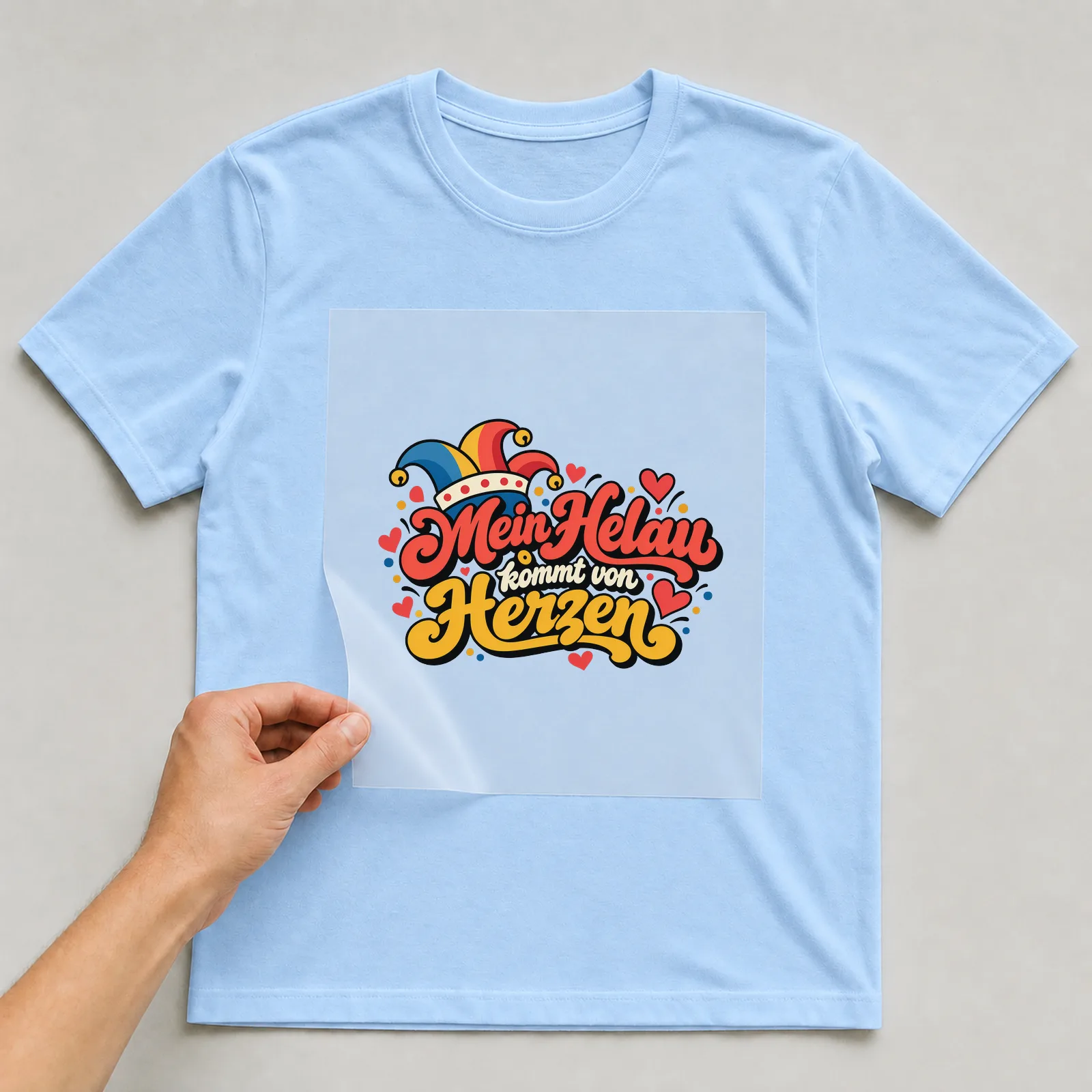 Bügelbild „Mein Helau kommt von Herzen“ auf hellblauem T-Shirt, Folie wird aufgelegt