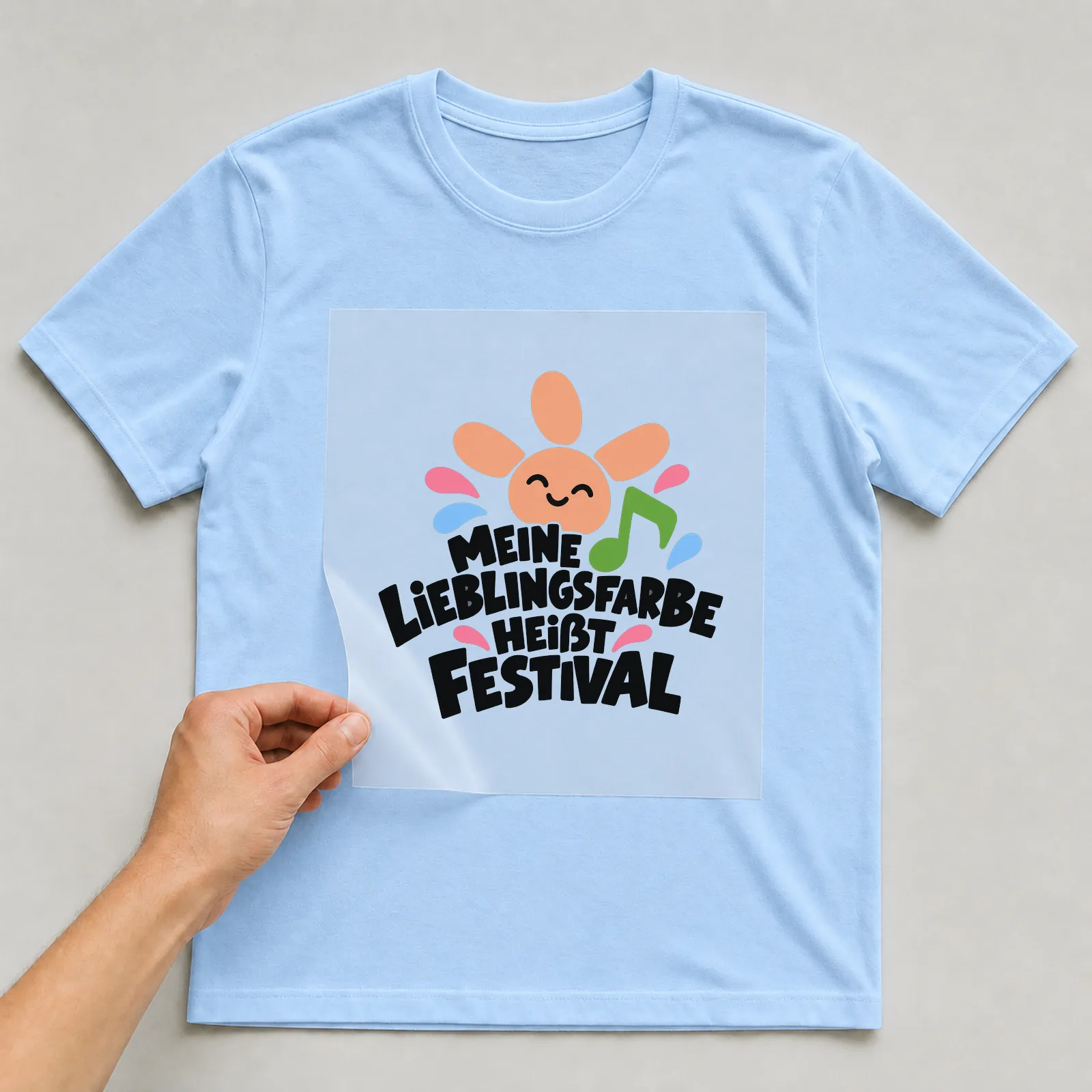 Bügelbild „MEINE LIEBLINGSFARBE HEIßT FESTIVAL“ auf hellblauem T-Shirt, Folie wird aufgelegt