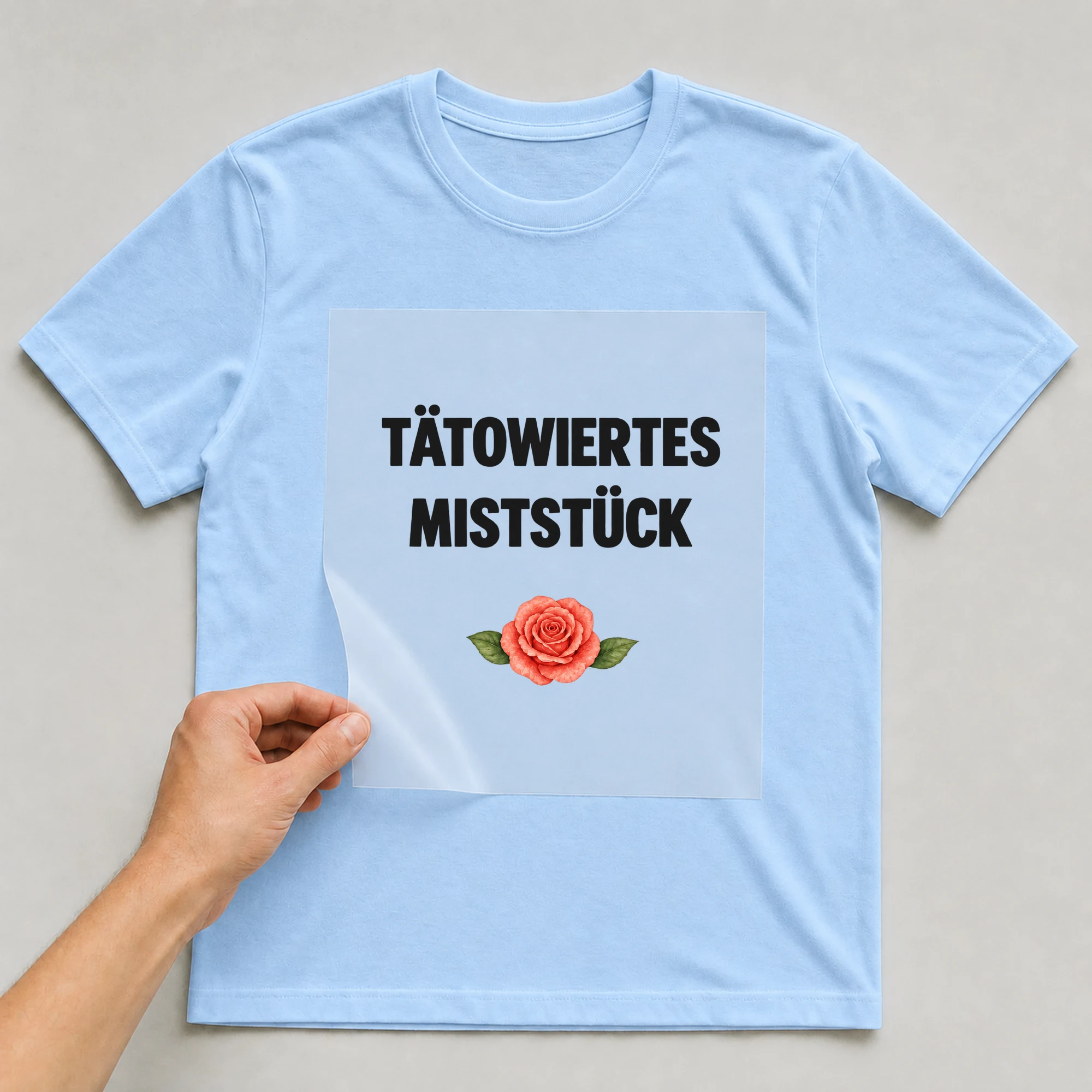 Bügelbild „TÄTOWIERTES MISTSTÜCK“ auf babyblauem T-Shirt, Folie wird aufgelegt