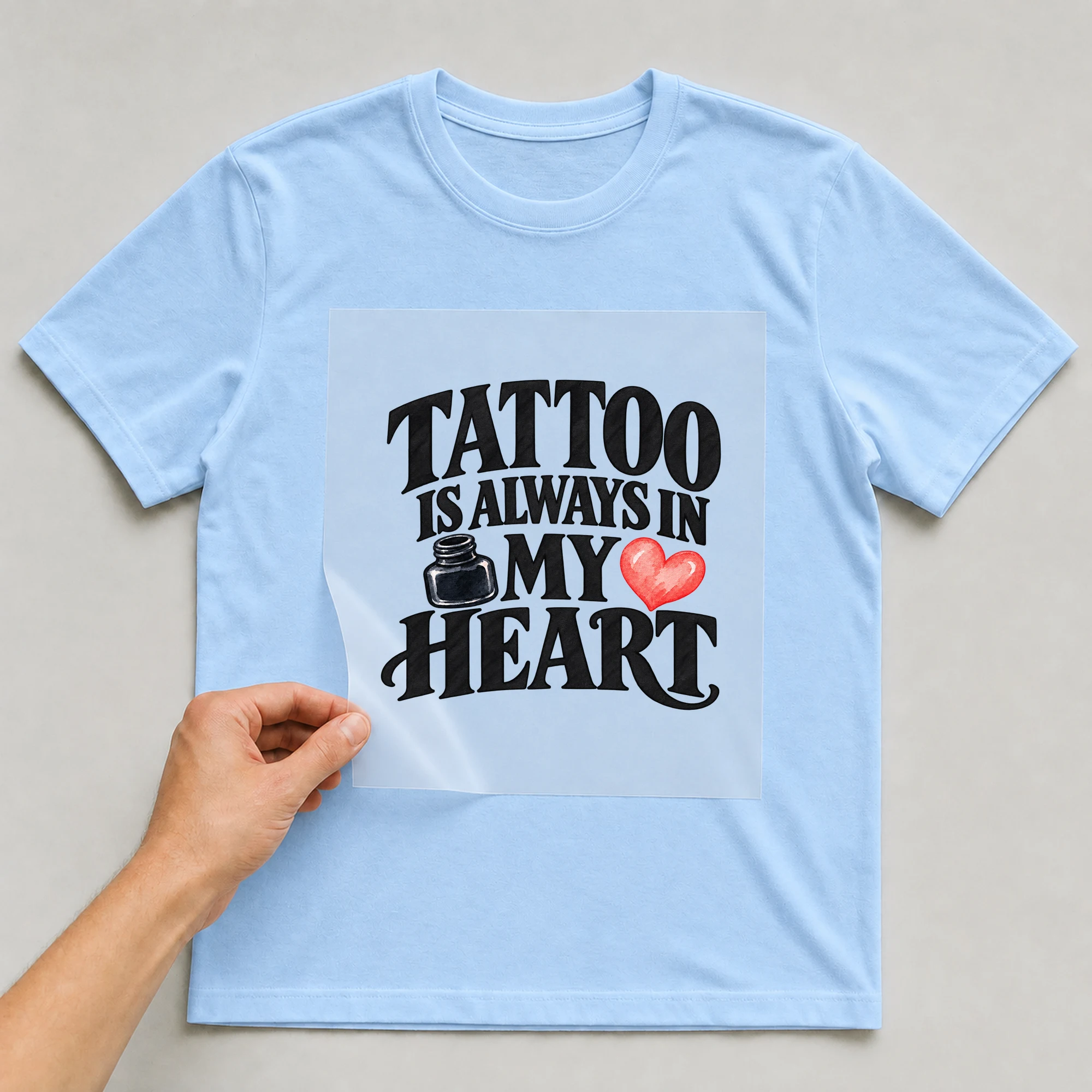 Bügelbild „Tattoo Is Always In My Heart“ auf babyblauem T-Shirt, Folie wird aufgelegt