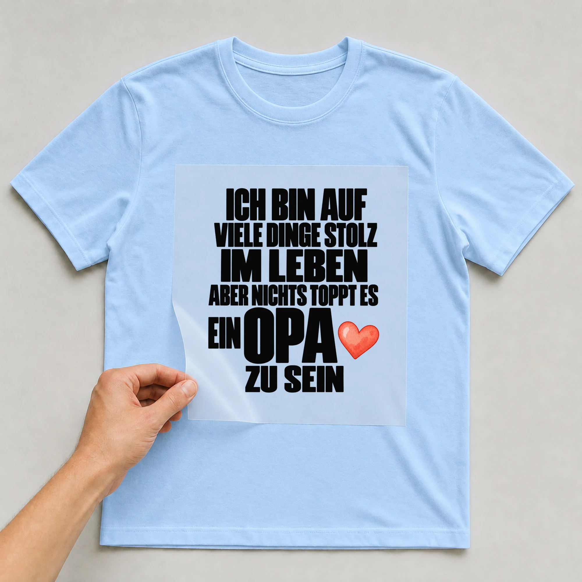 Bügelbild „Ich bin auf viele Dinge stolz im Leben aber nichts toppt es ein Opa zu sein“ auf babyblauem T-Shirt, Folie wird aufgelegt