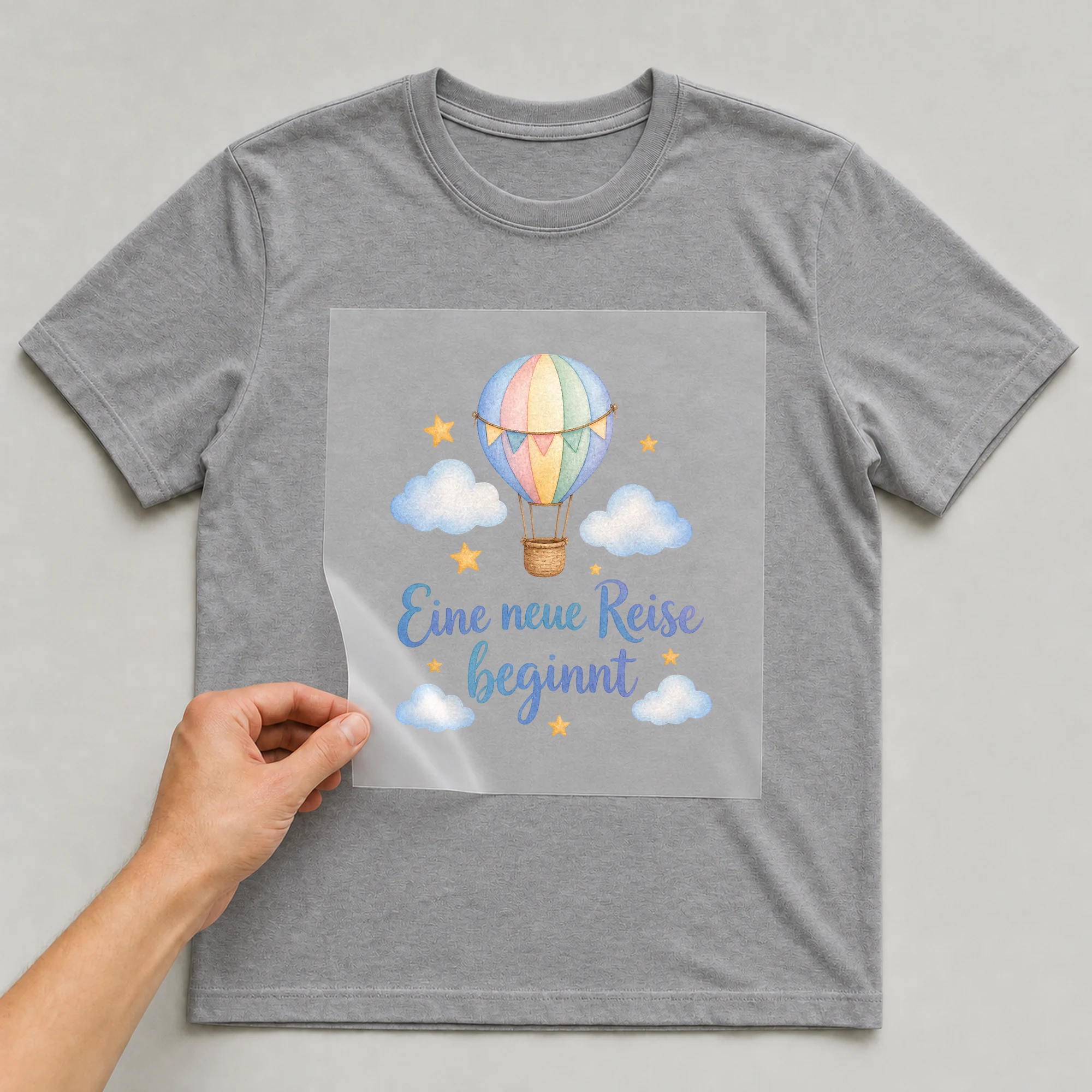 Bügelbild „Eine neue Reise beginnt“ auf grauem T-Shirt, Folie wird aufgelegt