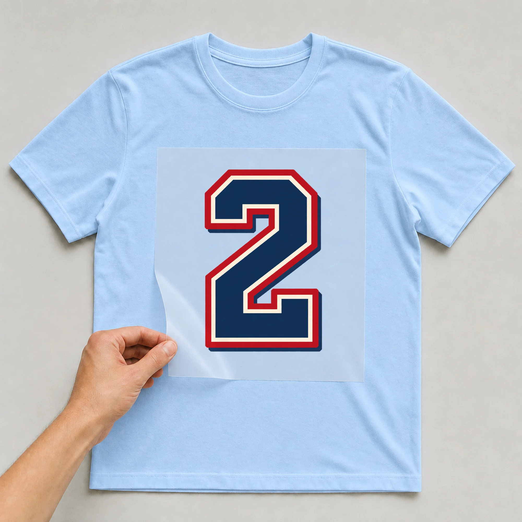 Bügelbild „Zahl 2 im Retro-College-Stil“ auf babyblauem T-Shirt, Folie wird aufgelegt