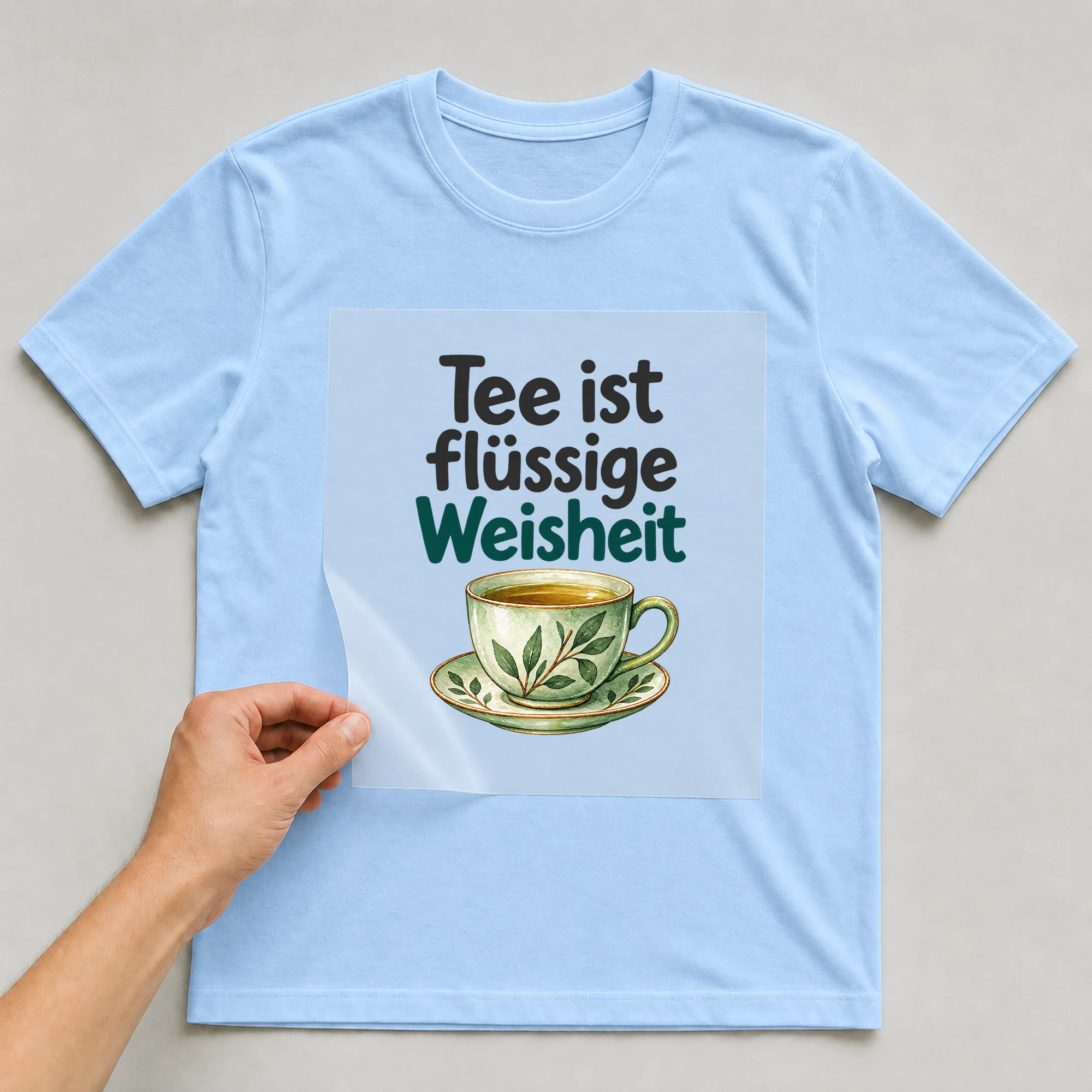 Bügelbild „Tee ist flüssige Weisheit“ auf babyblauem T-Shirt, Folie wird aufgelegt