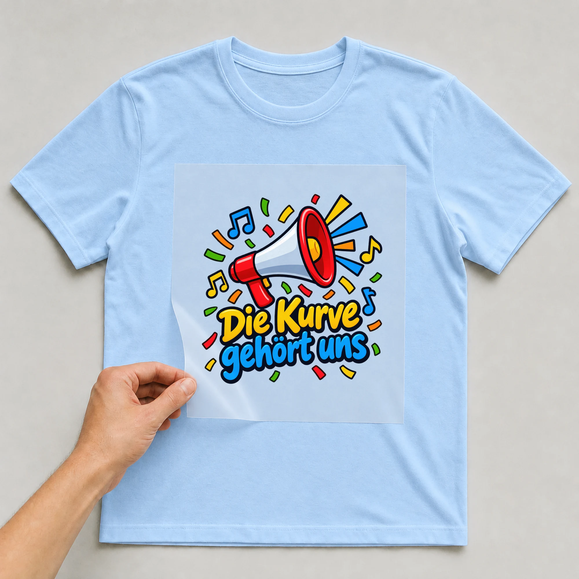 Bügelbild „Die Kurve gehört uns“ auf babyblauem T-Shirt, Folie wird aufgelegt