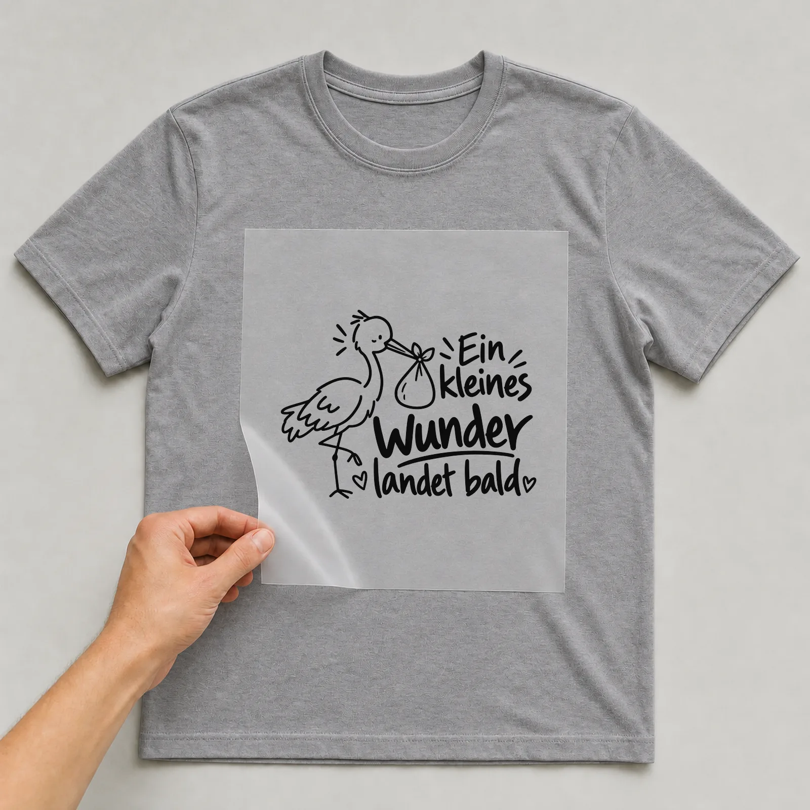 Bügelbild „Ein kleines Wunder landet bald“ auf grauem T-Shirt, Folie wird aufgelegt