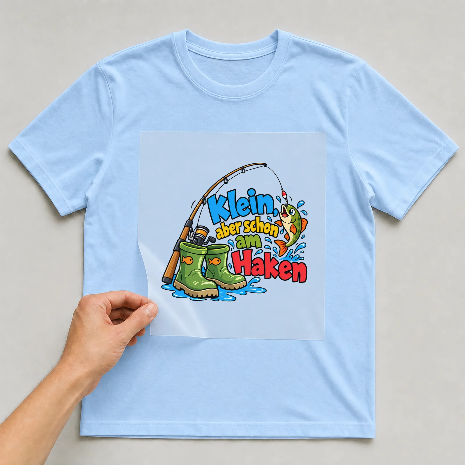 Bügelbild „Klein, aber schon am Haken“ auf hellblauem T-Shirt, Folie wird aufgelegt