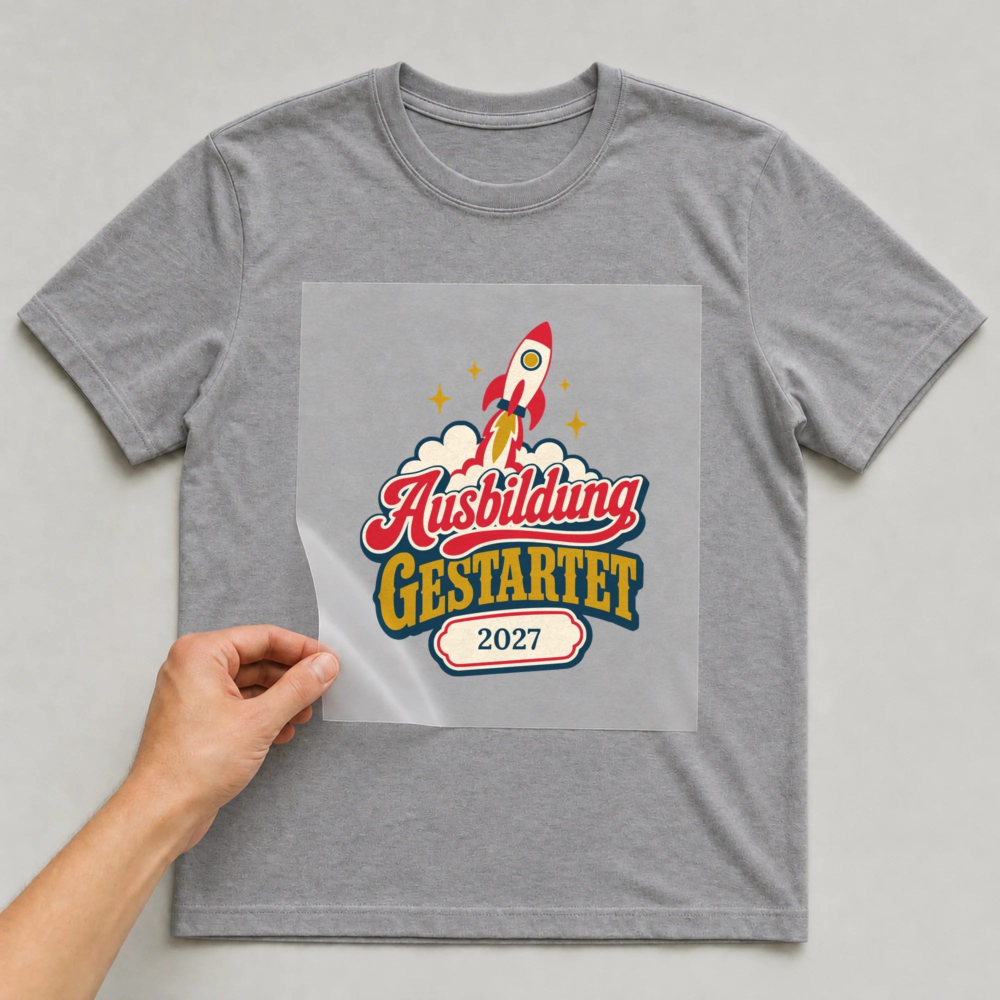 Bügelbild Ausbildung gestartet mit Jahr auf grauem T-Shirt, Folie wird aufgelegt