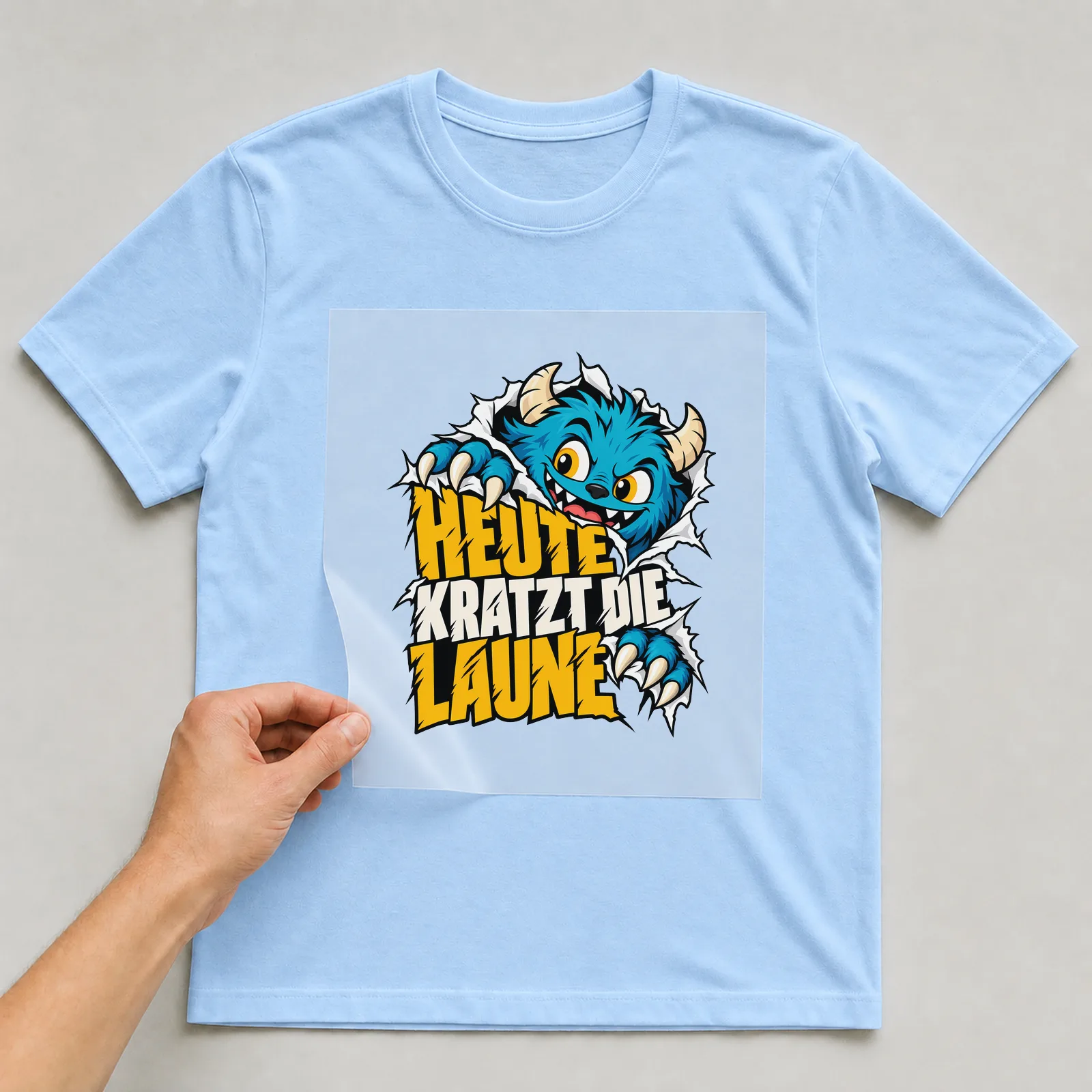 Bügelbild „Heute kratzt die Laune“ auf hellblauem T-Shirt, Folie wird aufgelegt