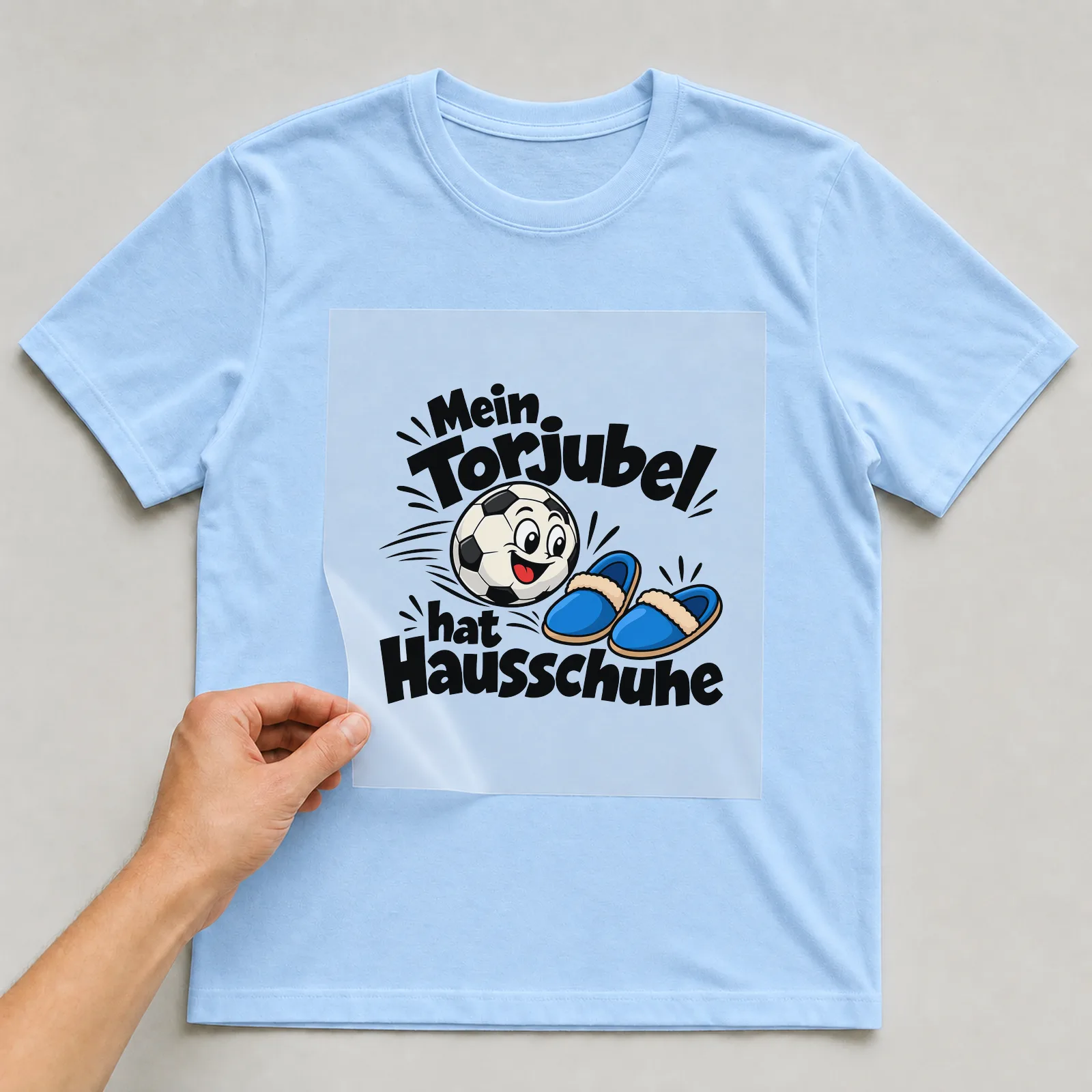 Bügelbild „Mein Torjubel hat Hausschuhe“ auf hellblauem T-Shirt, Folie wird aufgelegt