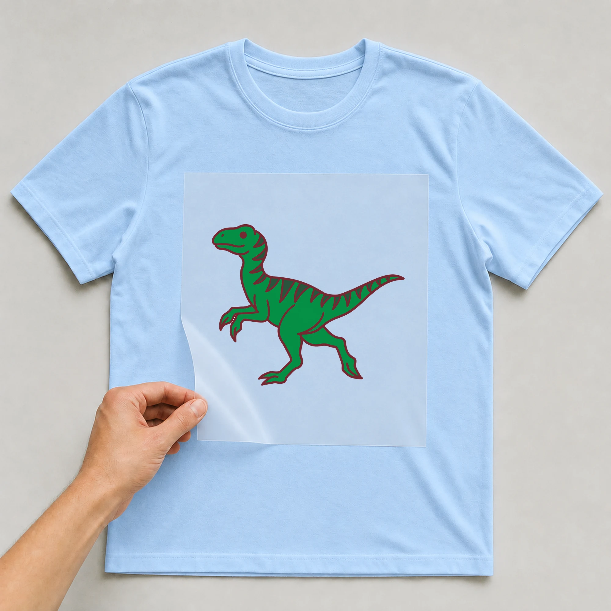 Bügelbild „Dino Raptor“ auf babyblauem T-Shirt, Folie wird aufgelegt