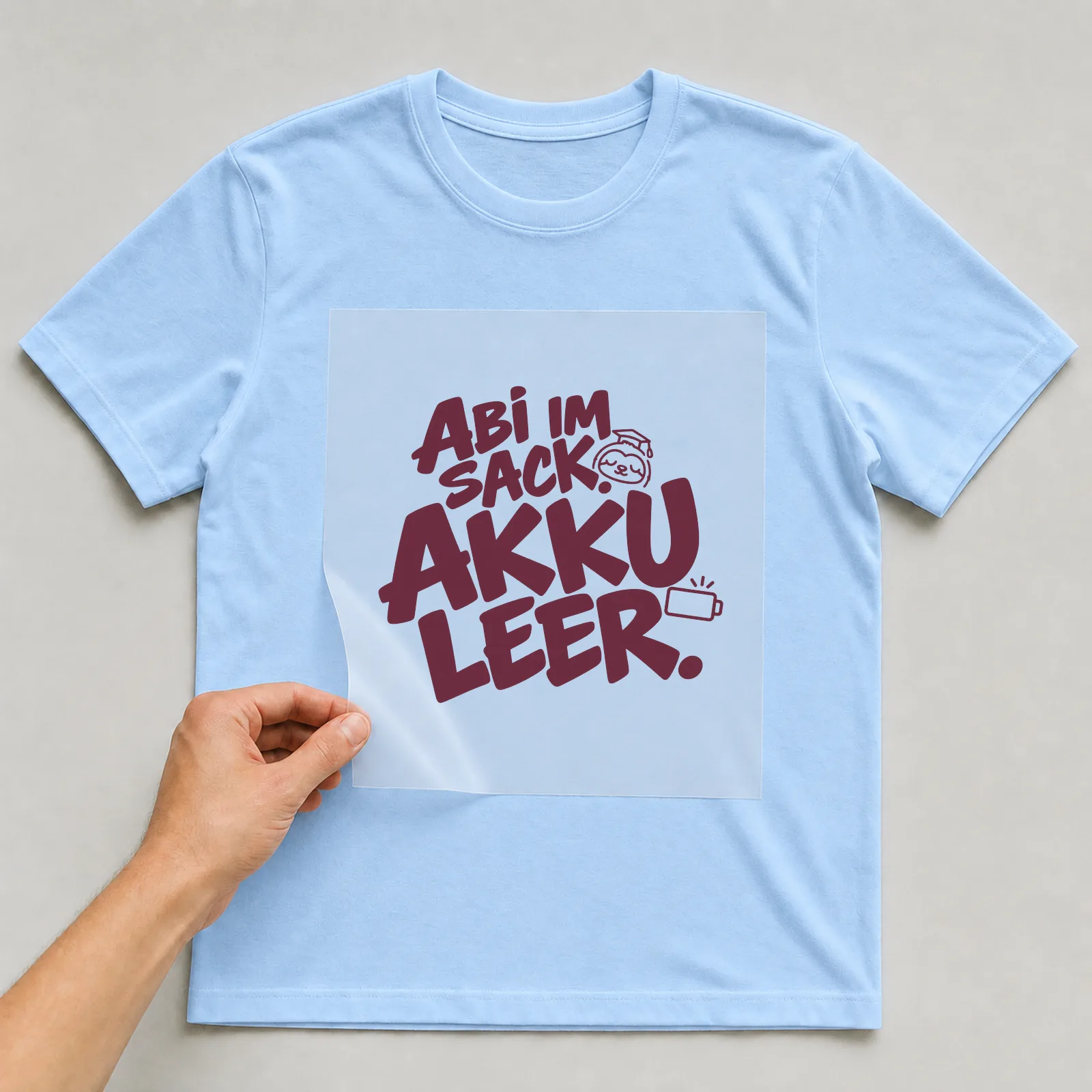Bügelbild „Abi im Sack. Akku leer.“ (Schrift Konturen, Nr. 2) auf hellblauem T-Shirt, Folie wird aufgelegt