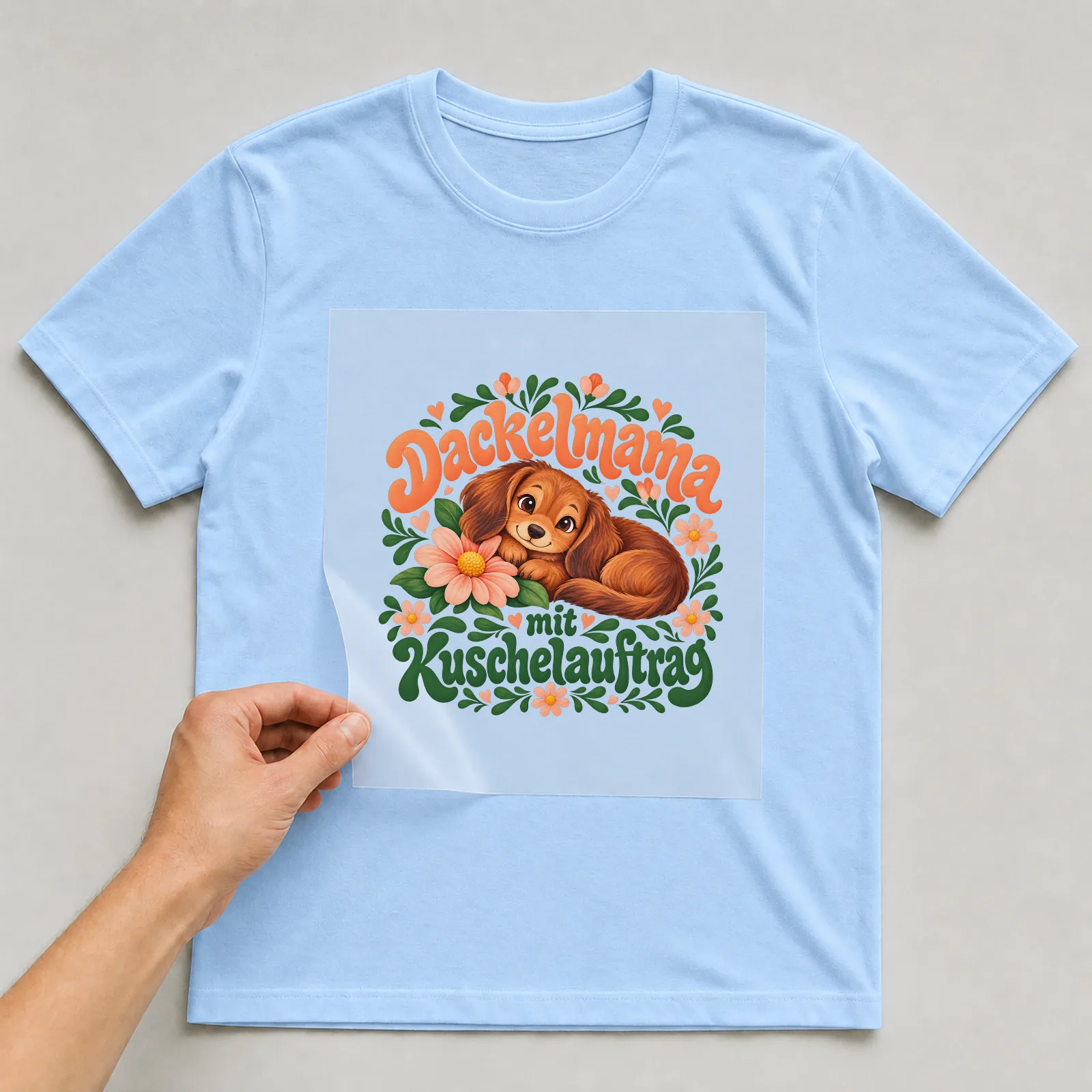 Bügelbild „Dackelmama mit Kuschelauftrag“ auf hellblauem T-Shirt, Folie wird aufgelegt