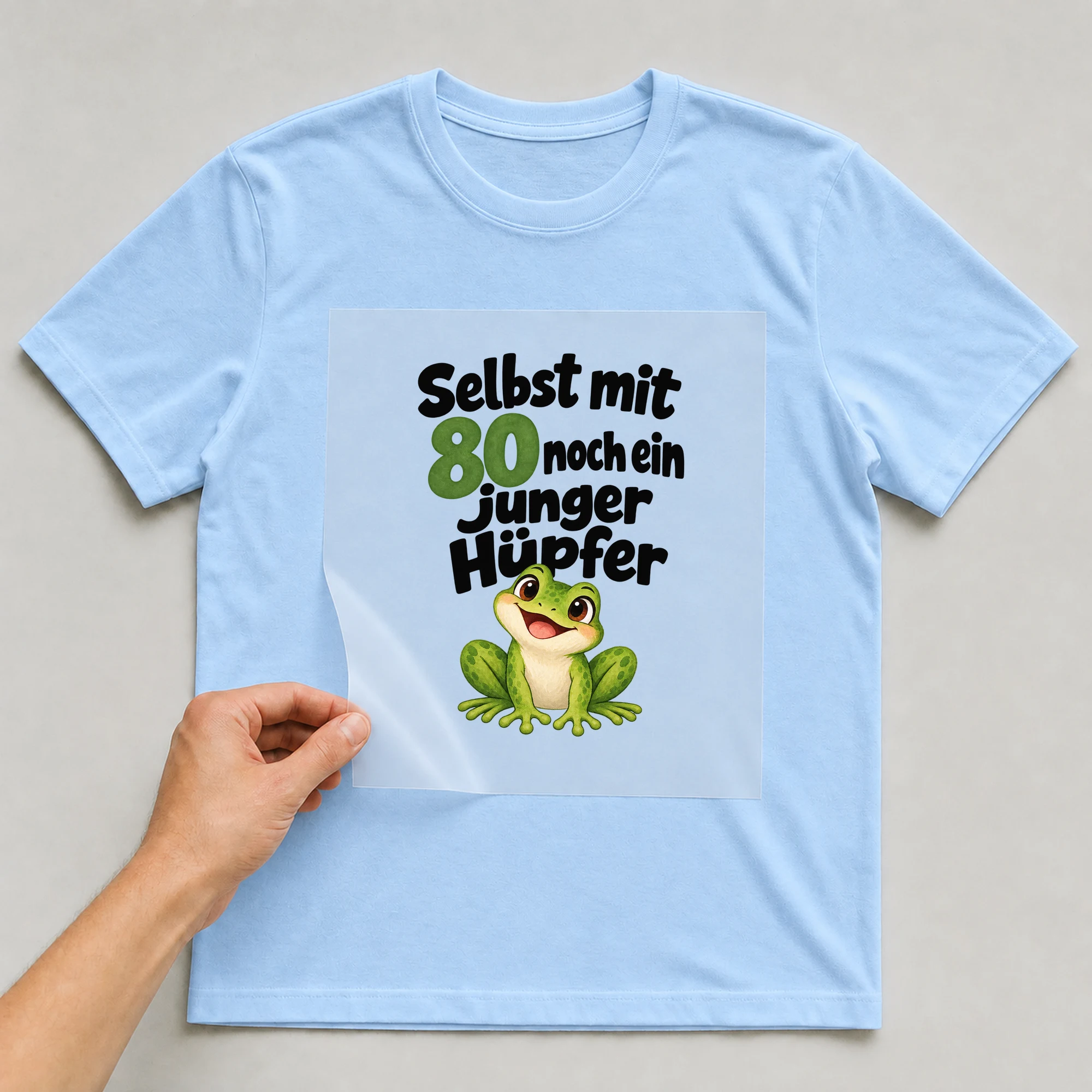 Bügelbild „Selbst mit 80 noch ein junger Hüpfer“ auf babyblauem T-Shirt, Folie wird aufgelegt