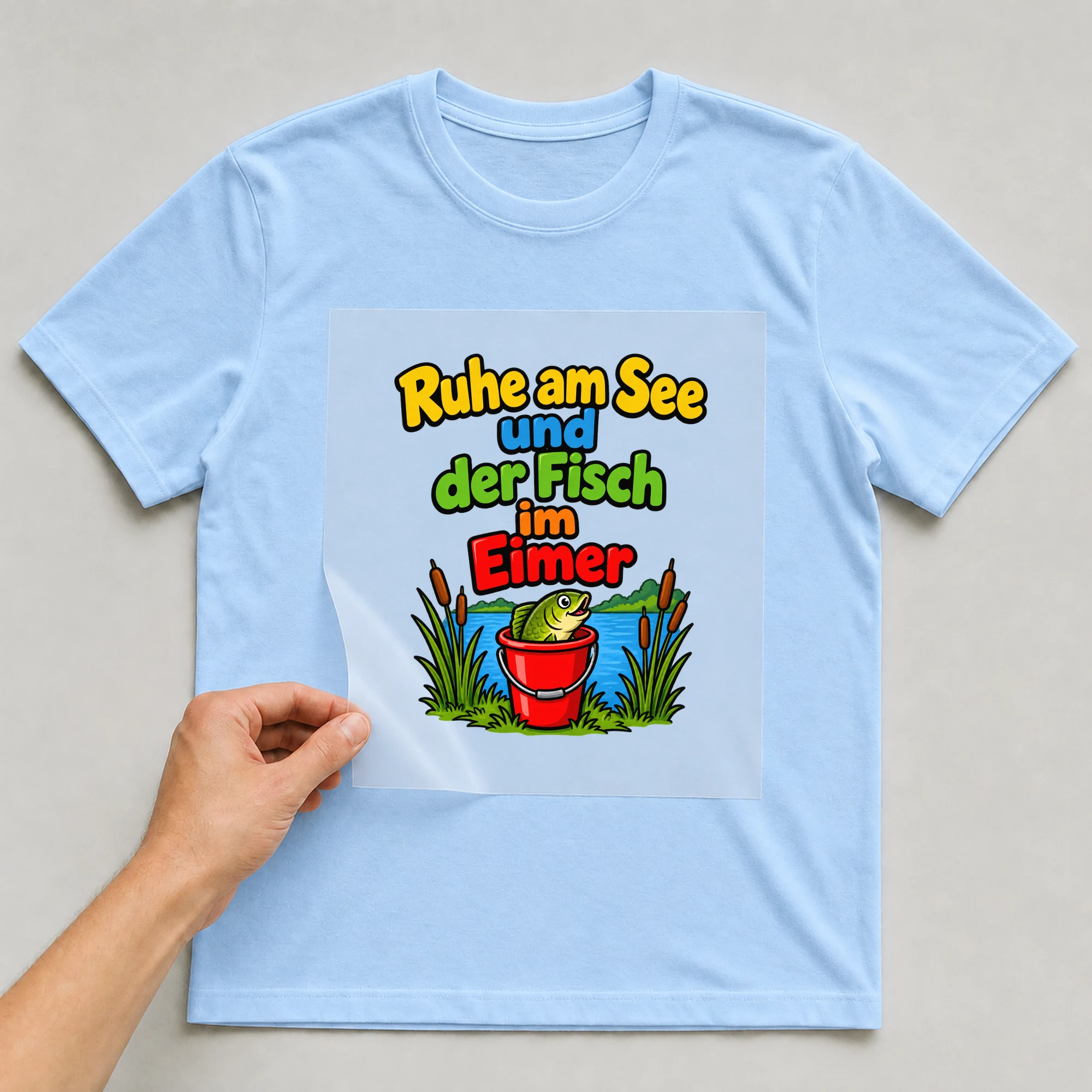 Bügelbild „Ruhe am See und der Fisch im Eimer“ auf babyblauem T-Shirt, Folie wird aufgelegt