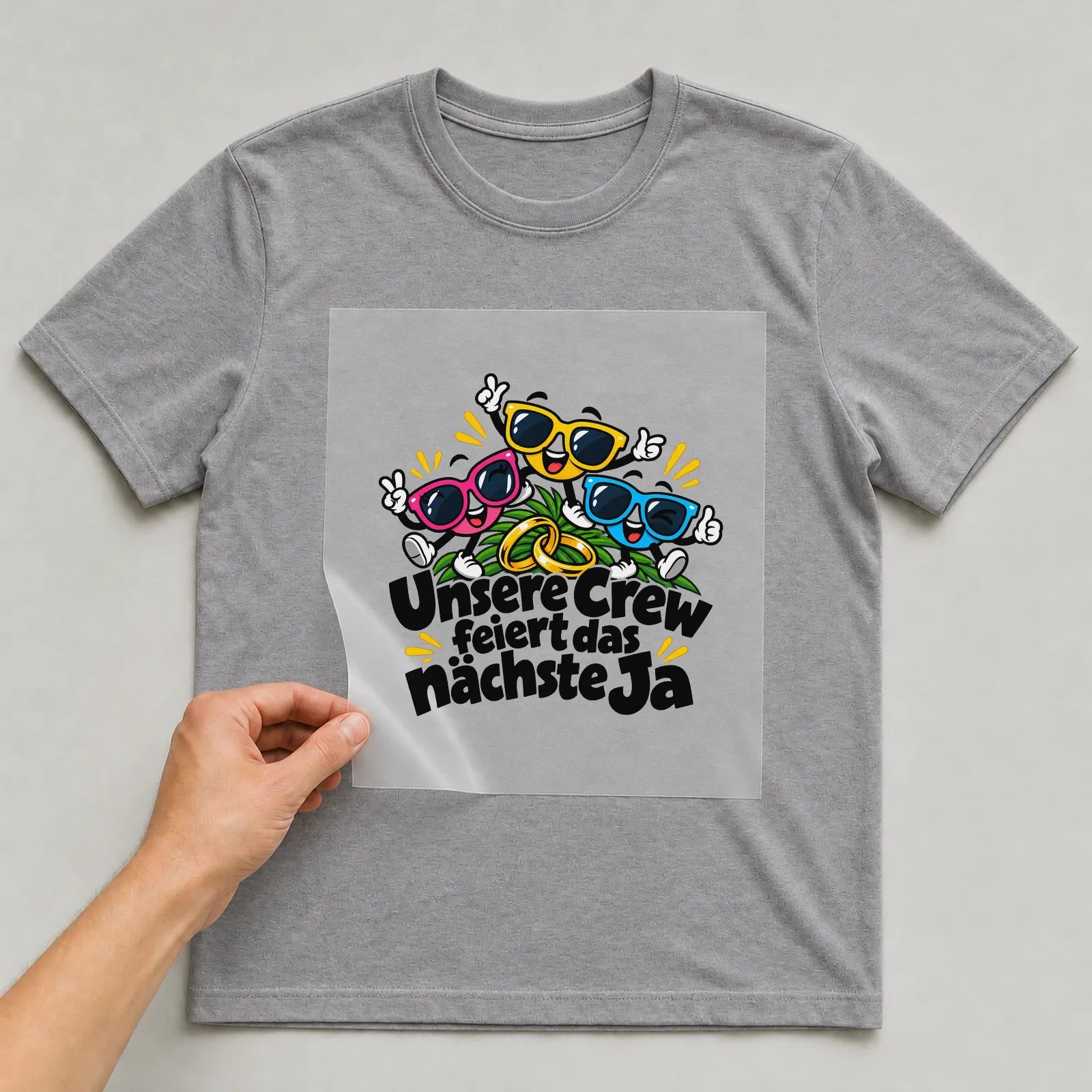 Bügelbild „Unsere Crew feiert das nächste Ja“ auf grauem T-Shirt, Folie wird aufgelegt