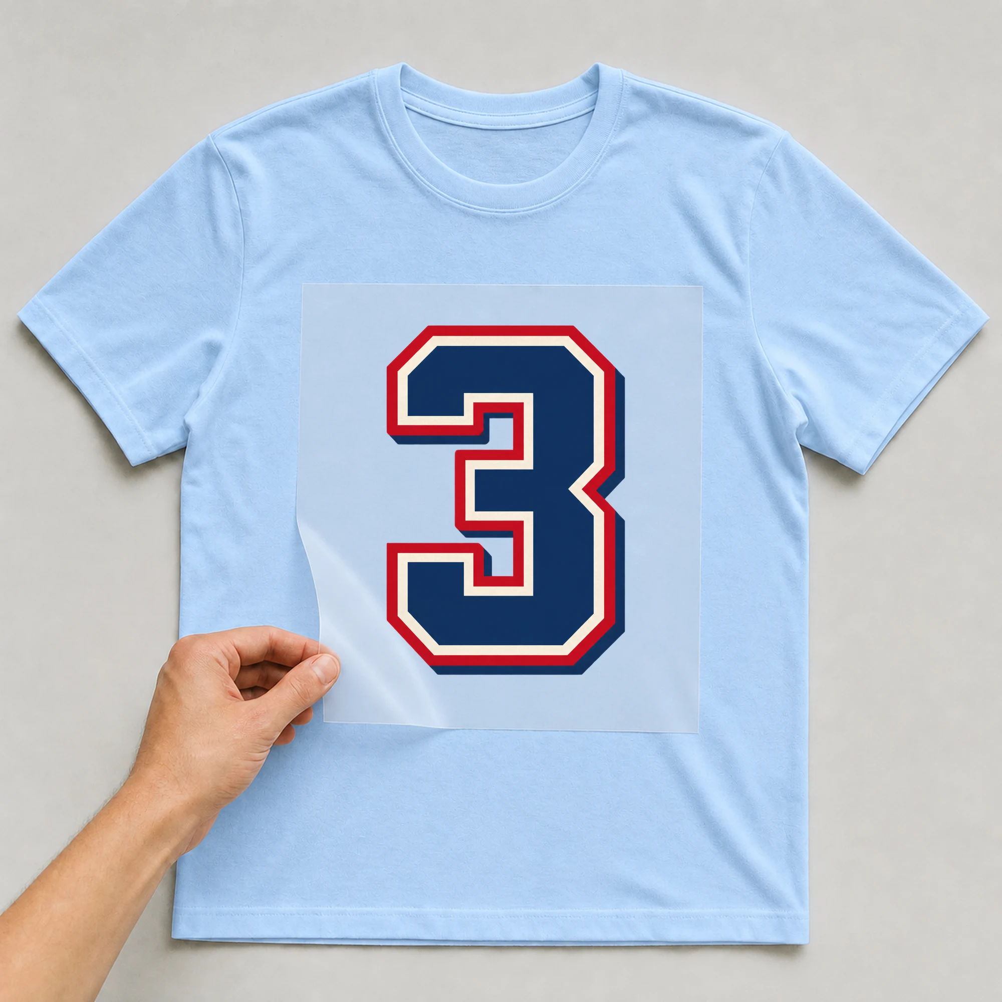 Bügelbild „Zahl 3 im Retro-College-Stil“ auf babyblauem T-Shirt, Folie wird aufgelegt