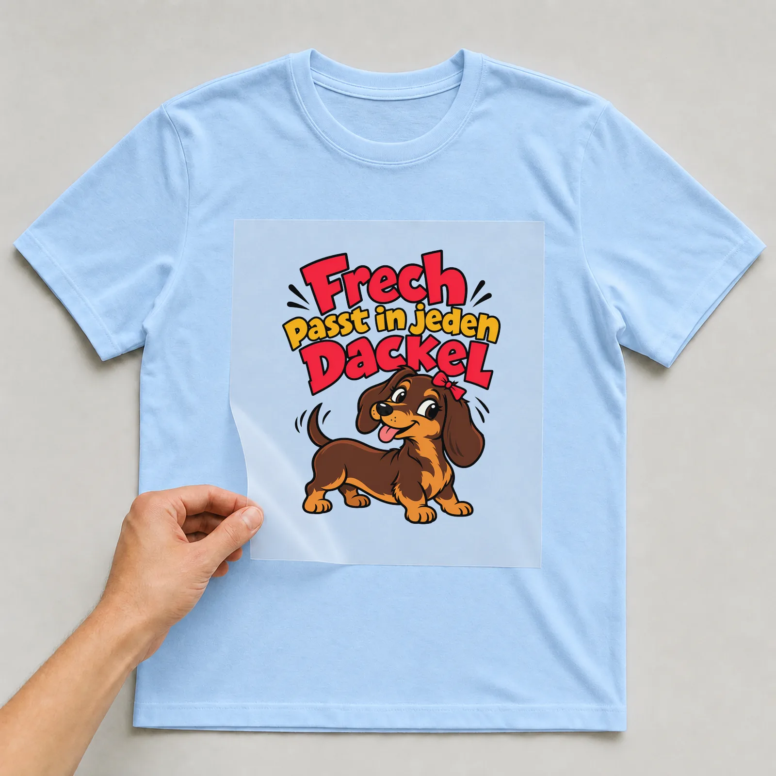 Bügelbild „Frech passt in jeden Dackel“ auf hellblauem T-Shirt, Folie wird aufgelegt