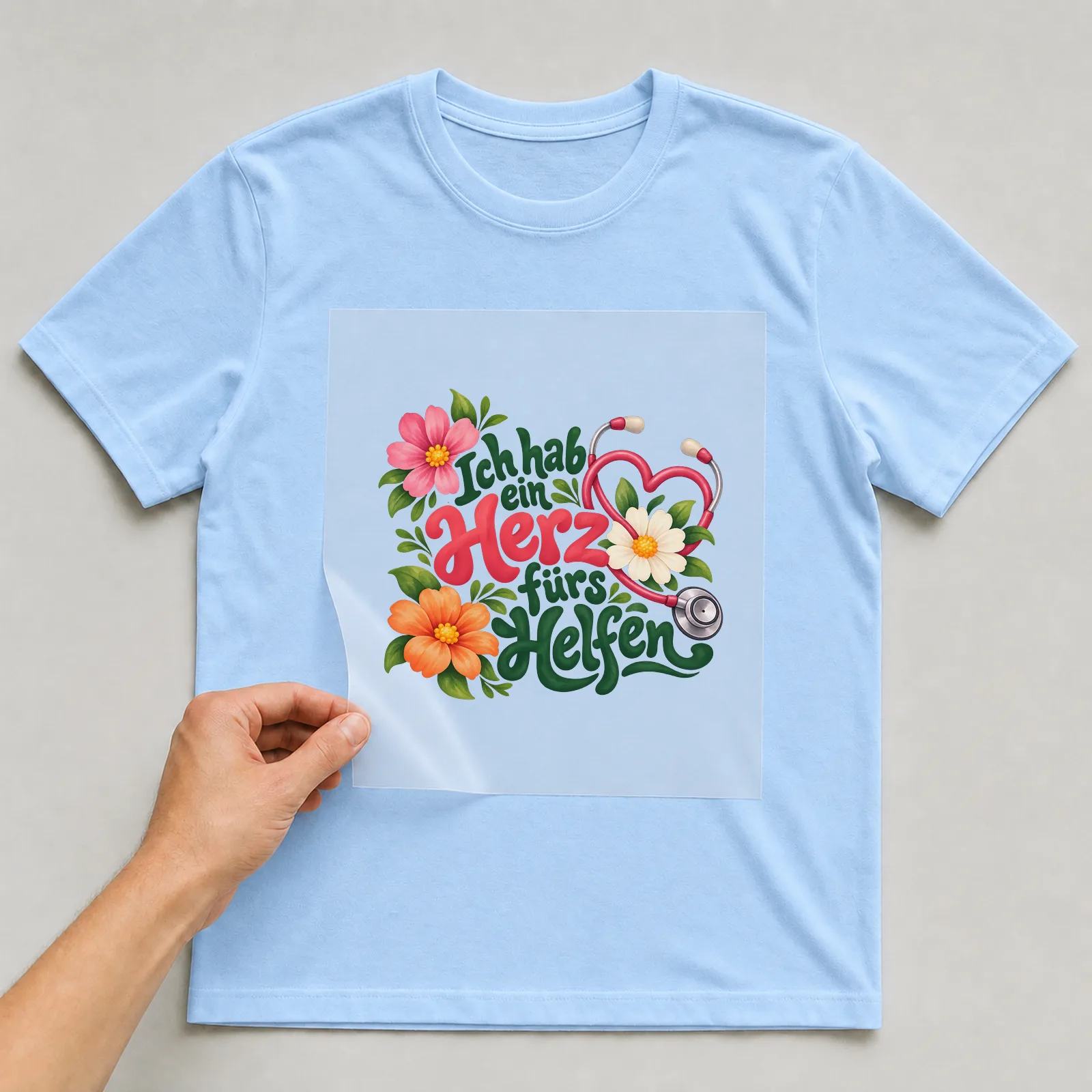 Bügelbild „Ich hab ein Herz fürs Helfen“ auf hellblauem T-Shirt, Folie wird aufgelegt