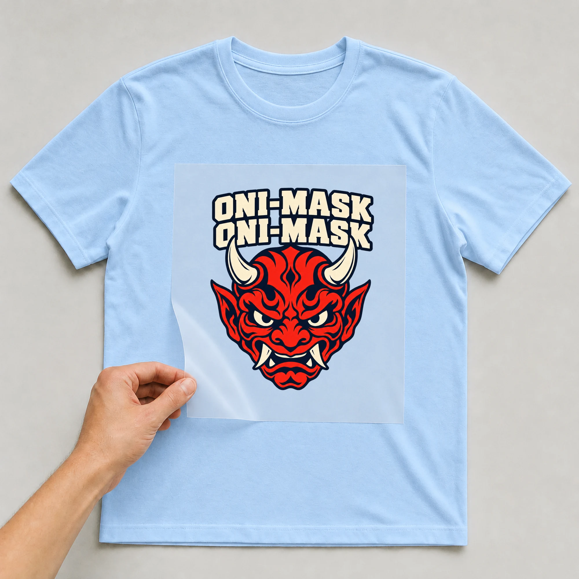 Bügelbild „Oni-mask Oni-mask“ auf babyblauem T-Shirt, Folie wird aufgelegt