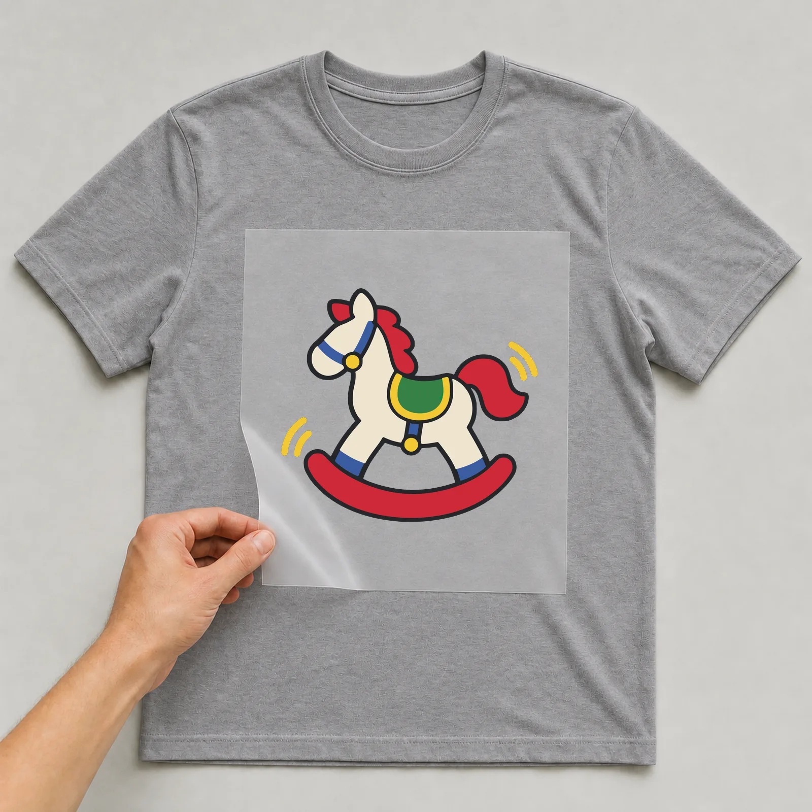 Bügelbild Schaukelpferd im Comic-Stil auf grauem T-Shirt, Folie wird aufgelegt