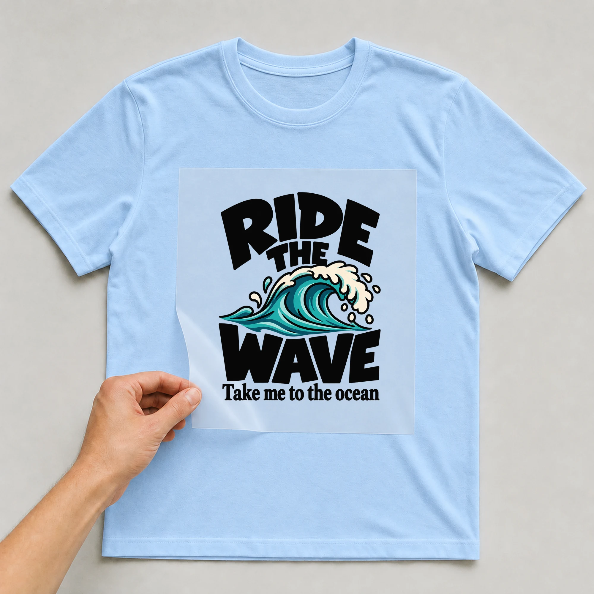 Bügelbild „Ride The Wave Take Me To The Ocean“ auf babyblauem T-Shirt, Folie wird aufgelegt