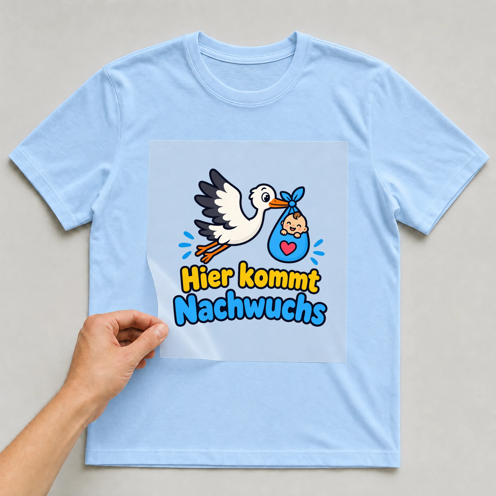 Bügelbild „Hier kommt Nachwuchs“ auf babyblauem T-Shirt, Folie wird aufgelegt