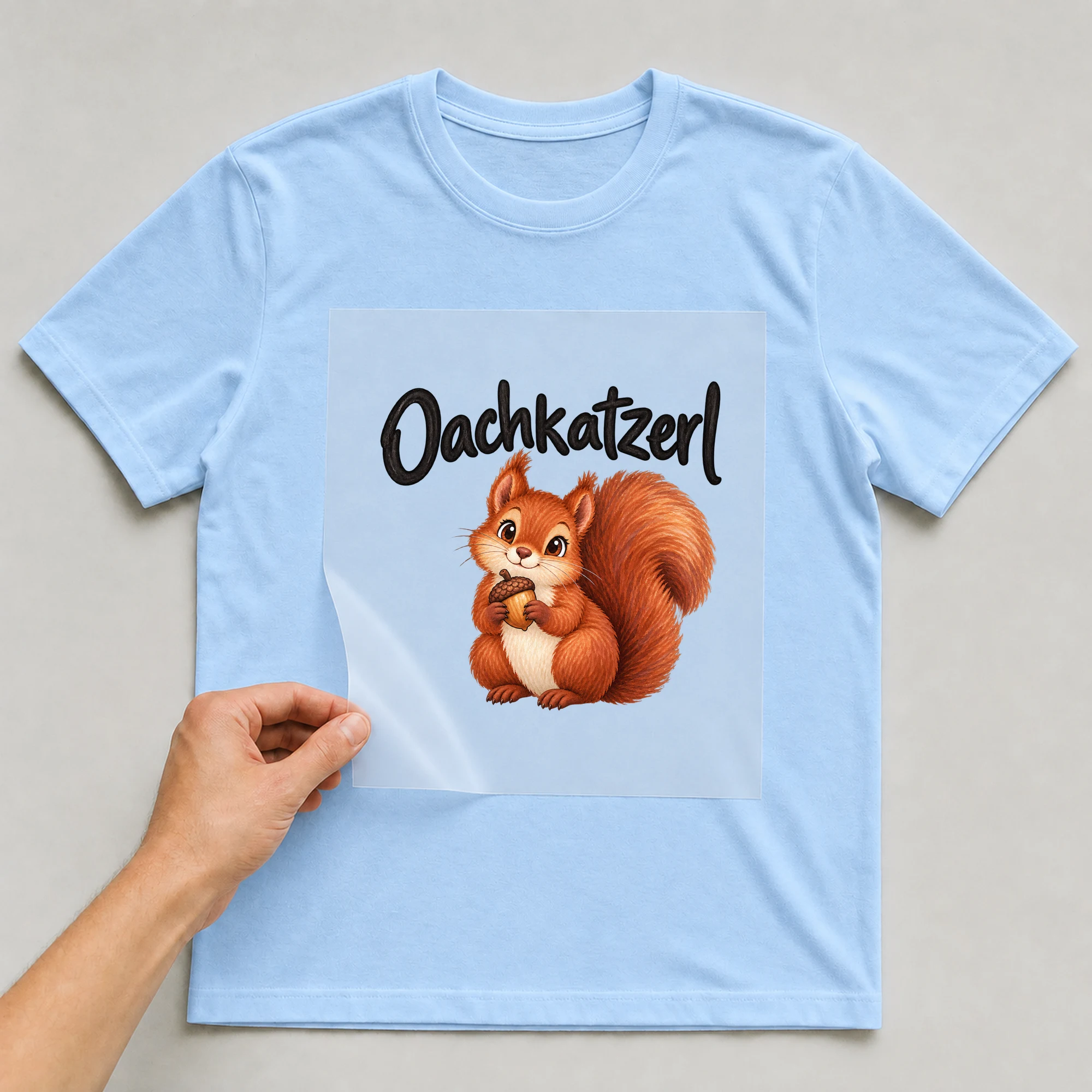 Bügelbild „Oachkatzerl“ auf babyblauem T-Shirt, Folie wird aufgelegt