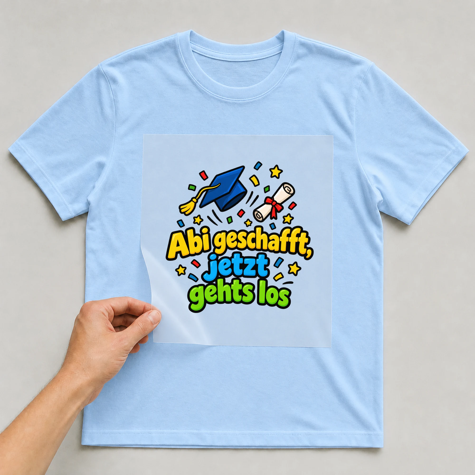 Bügelbild „Abi geschafft jetzt gehts los“ auf babyblauem T-Shirt, Folie wird aufgelegt