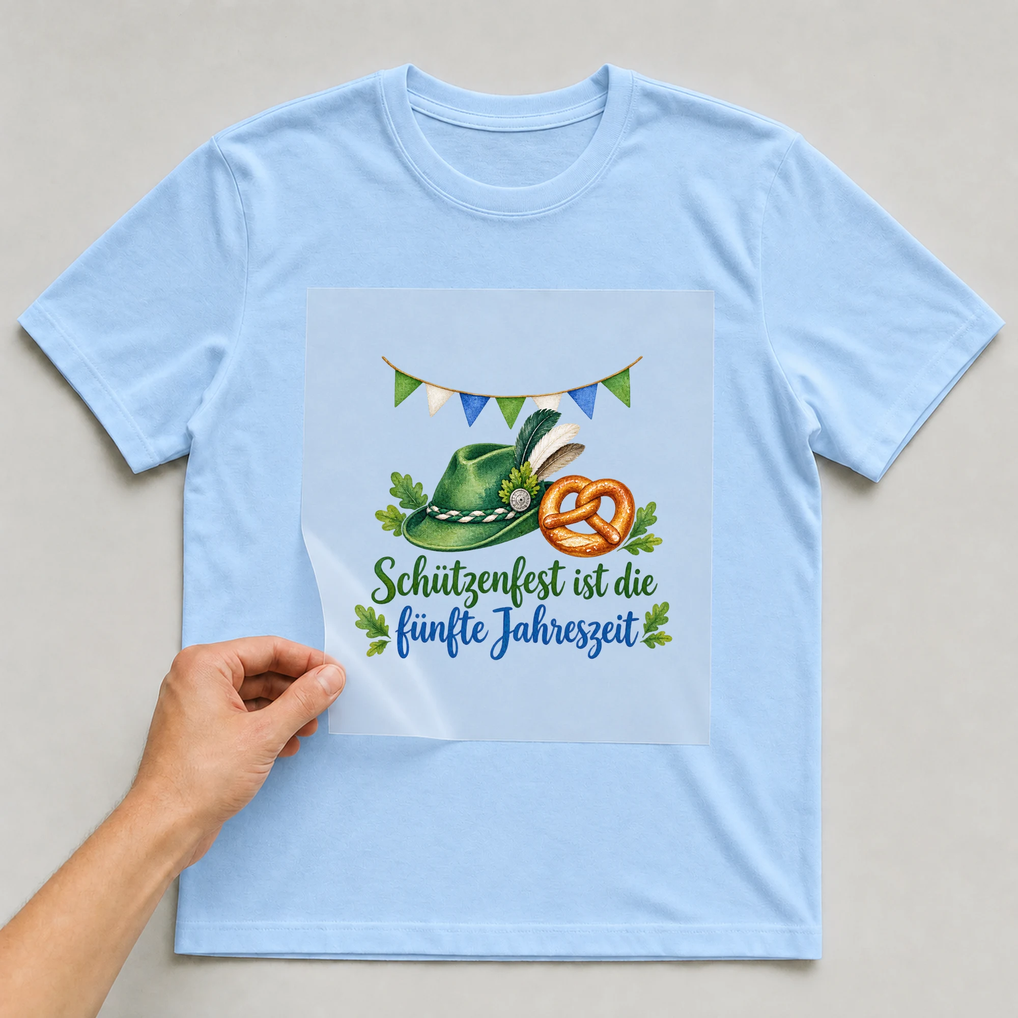 Bügelbild „Schützenfest ist die fünfte Jahreszeit“ auf babyblauem T-Shirt, Folie wird aufgelegt