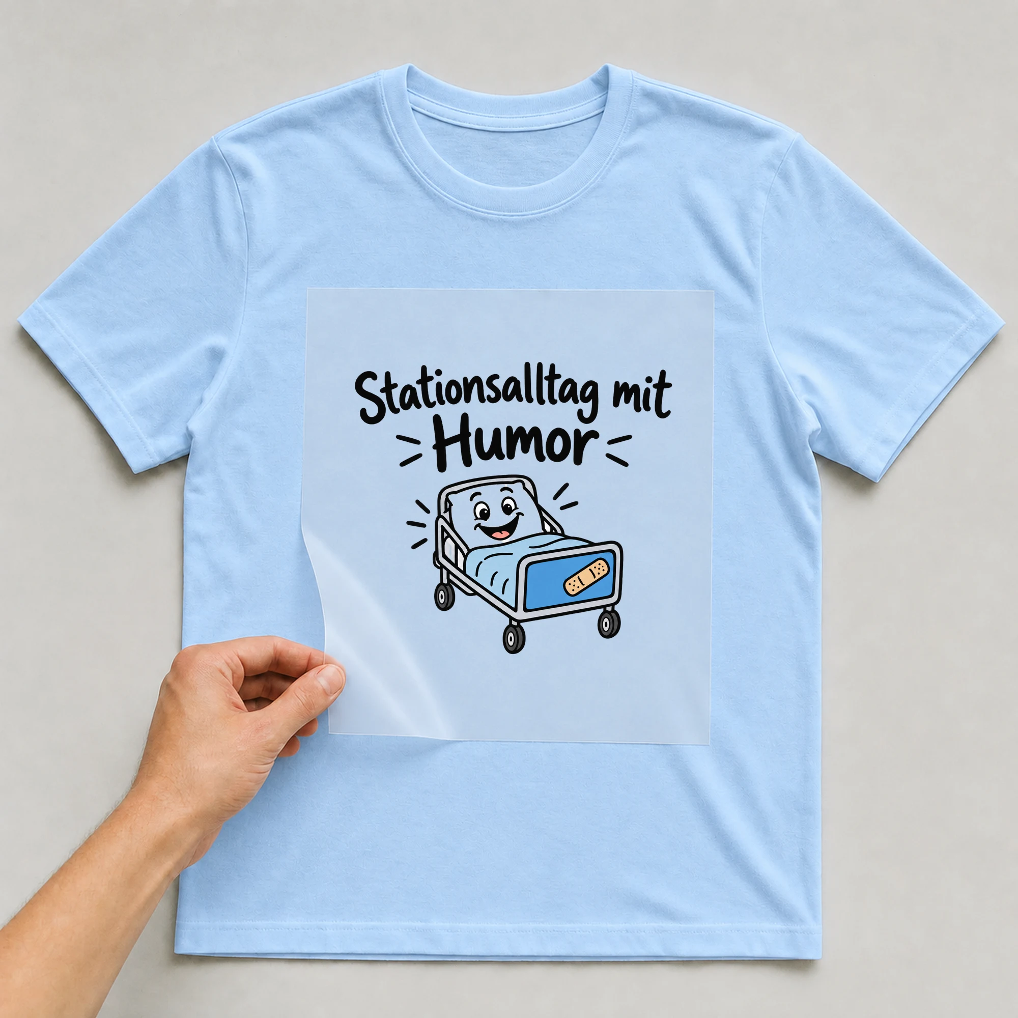 Bügelbild „Stationsalltag mit Humor“ auf babyblauem T-Shirt, Folie wird aufgelegt