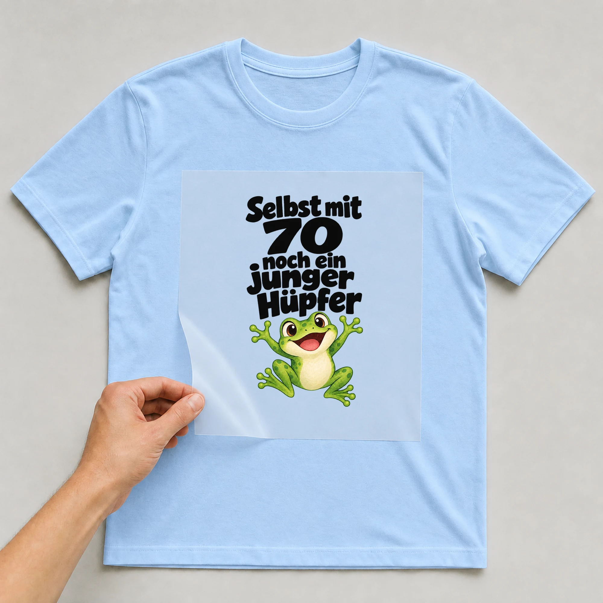 Bügelbild „Selbst mit 70 noch ein junger Hüpfer“ auf babyblauem T-Shirt, Folie wird aufgelegt