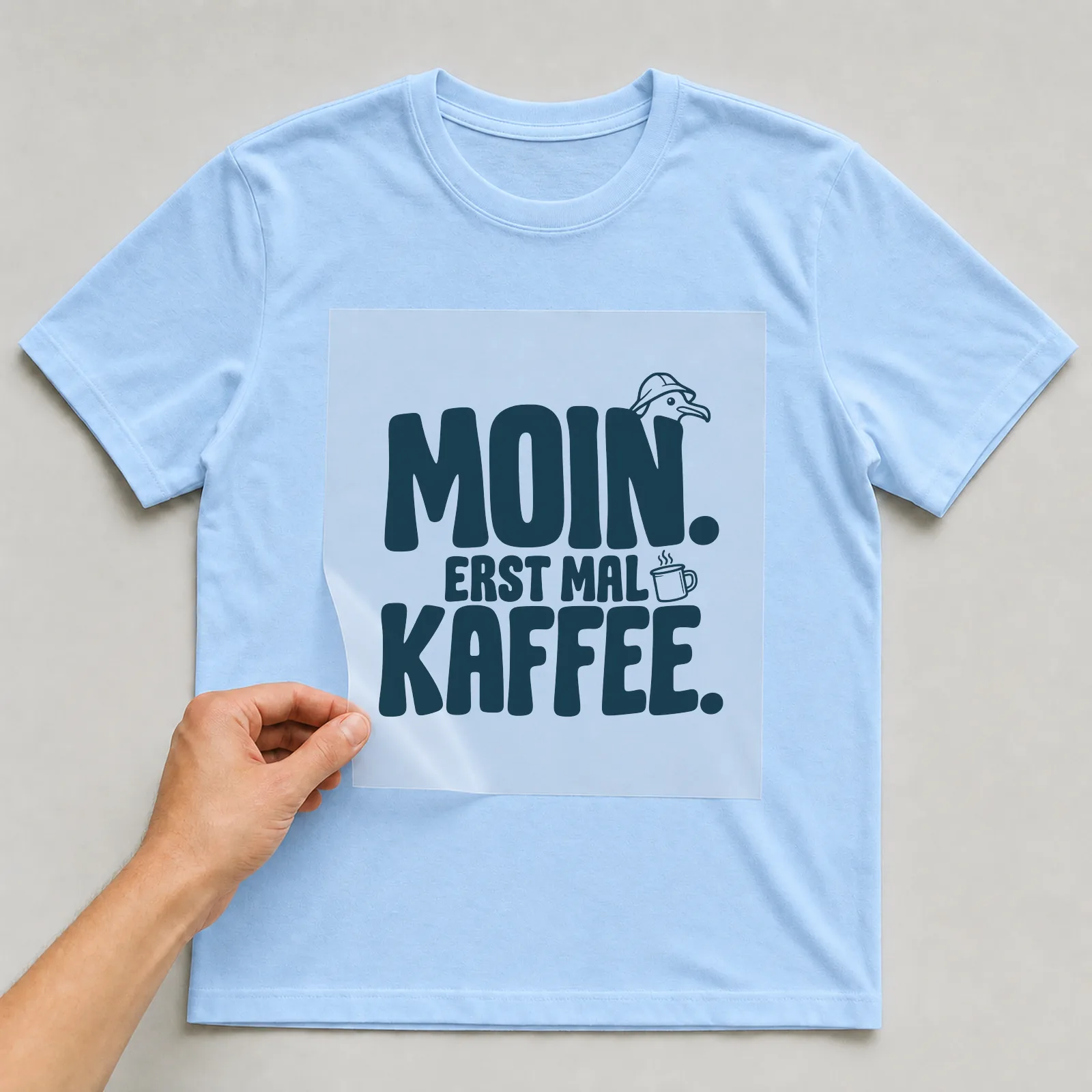 Bügelbild „Moin. Erst mal Kaffee.“ (Schrift Konturen, Nr. 3) auf hellblauem T-Shirt, Folie wird aufgelegt
